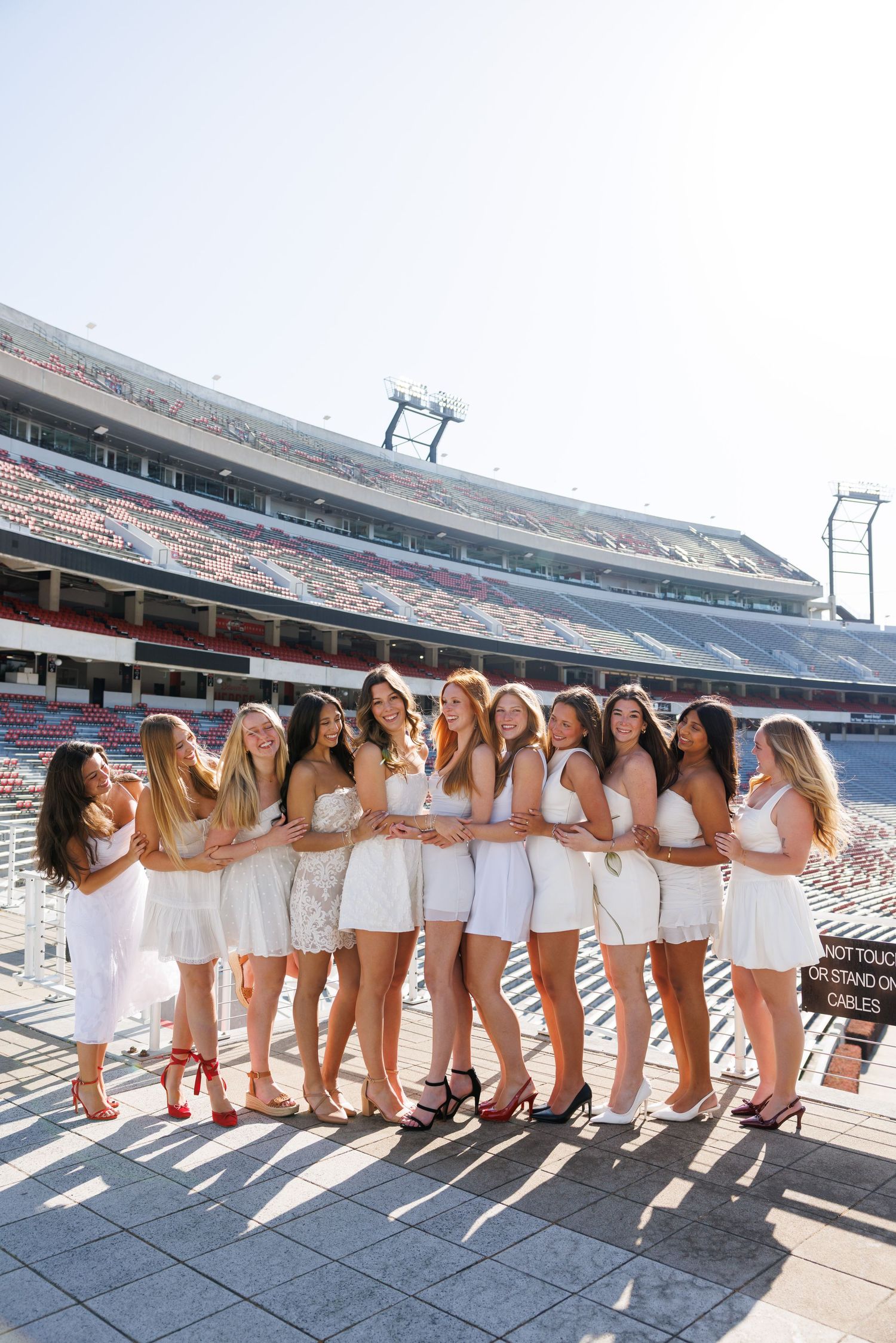 Group of women in white dresses posing together in a large stadium on a sunny day.