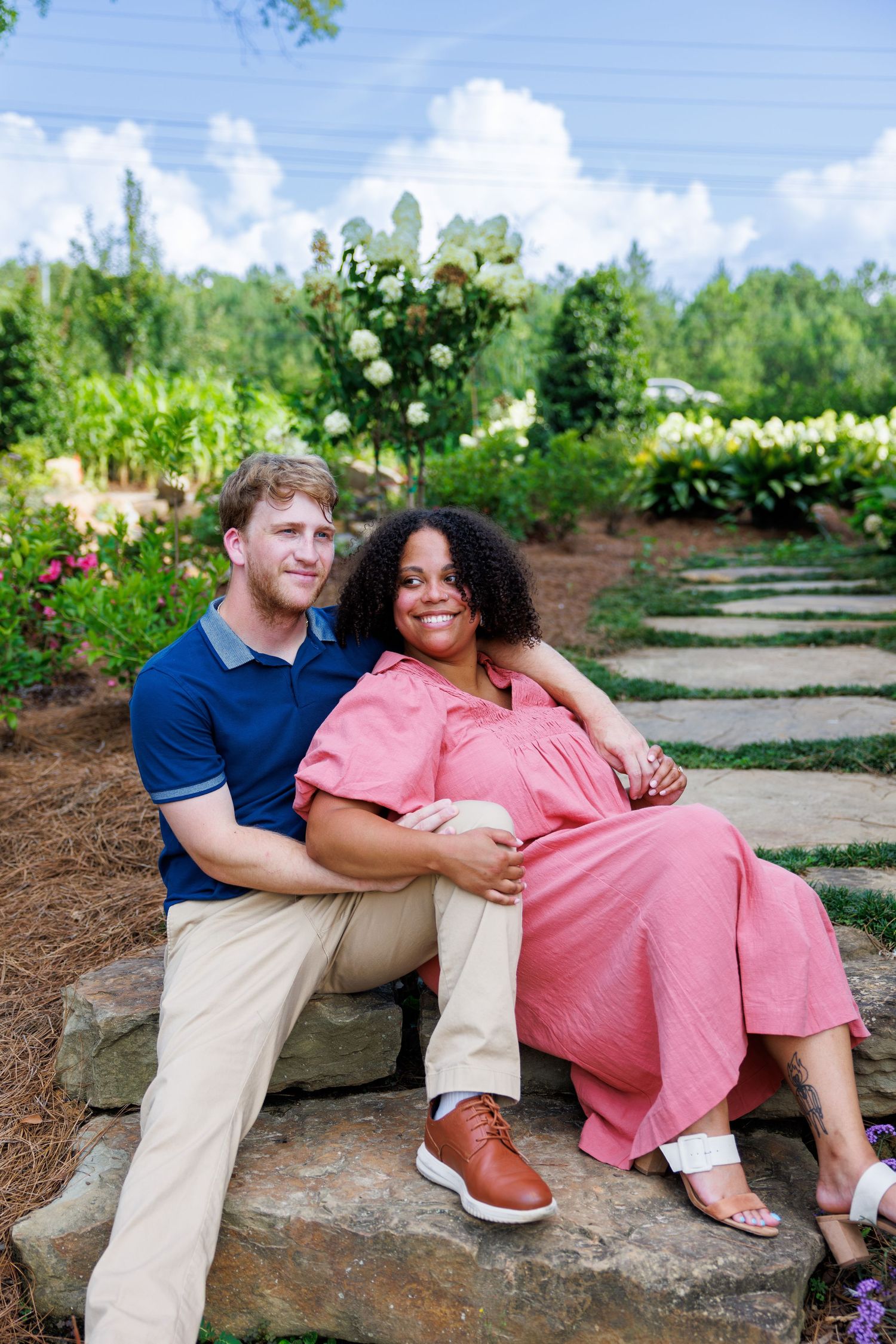 A couple sits on stone steps in a lush garden, the woman in a pink dress smiling warmly while her partner embraces her.