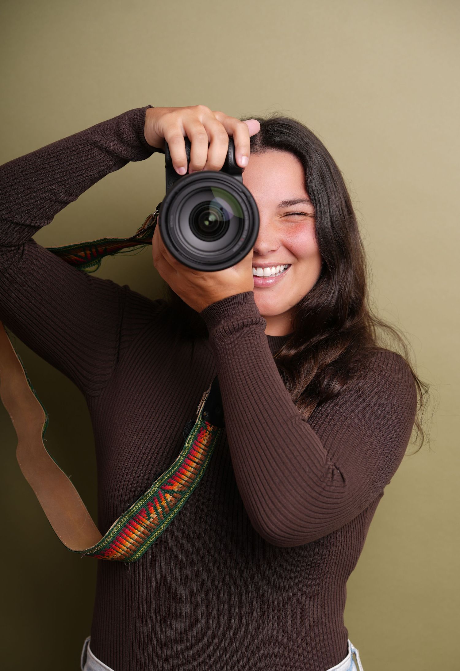 Photographer in a brown sweater holding a DSLR camera while taking pictures against a neutral background.