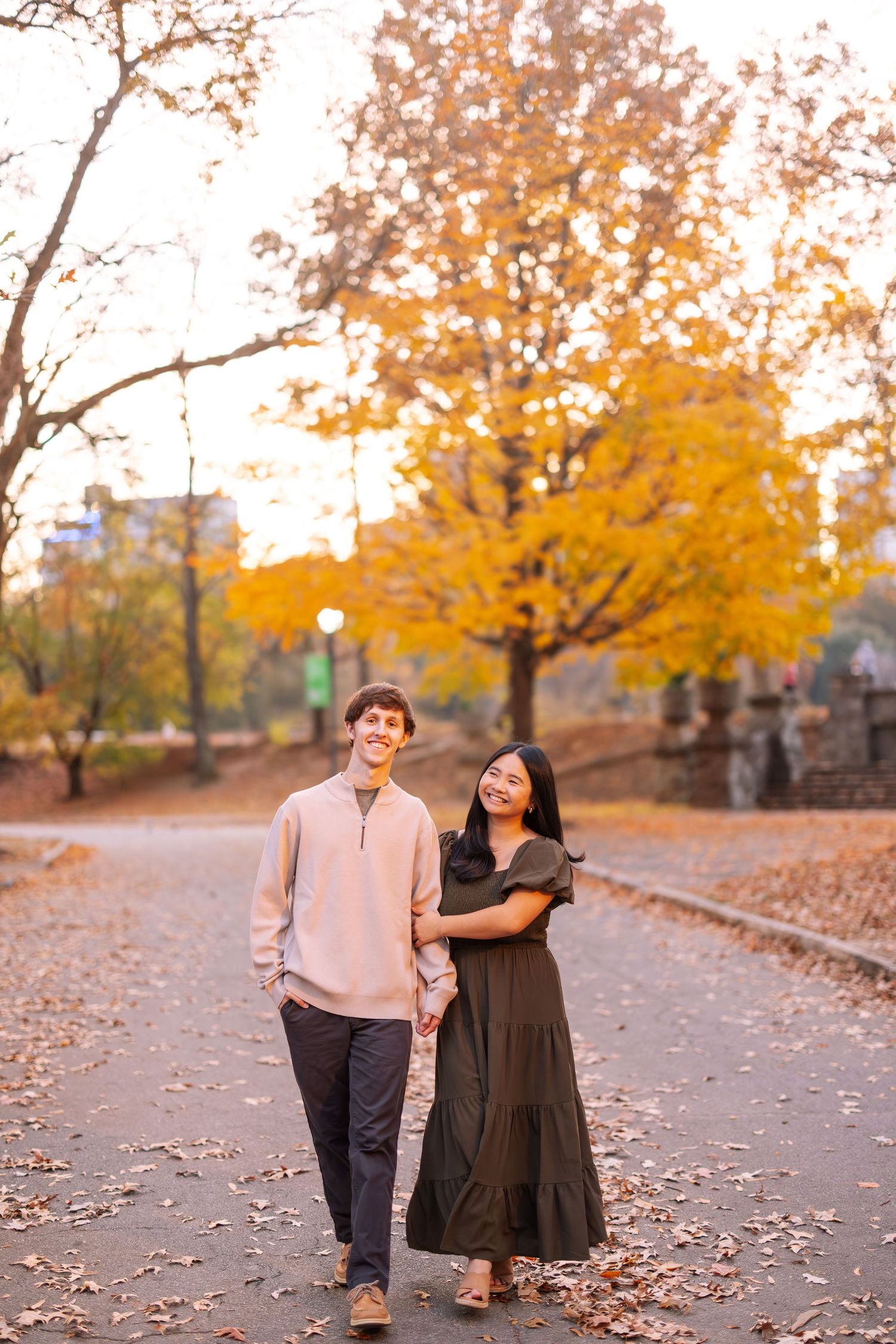 A couple walks together on a path lined with golden autumn trees in a park setting.
