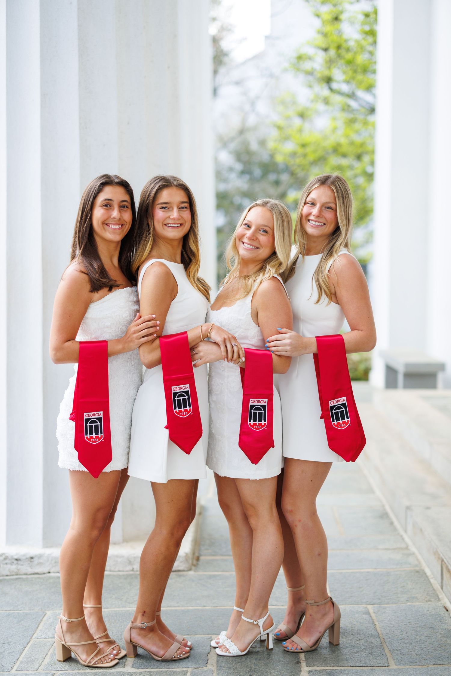 Four young women in white dresses holding red graduation stoles pose between white columns for a celebratory photo.