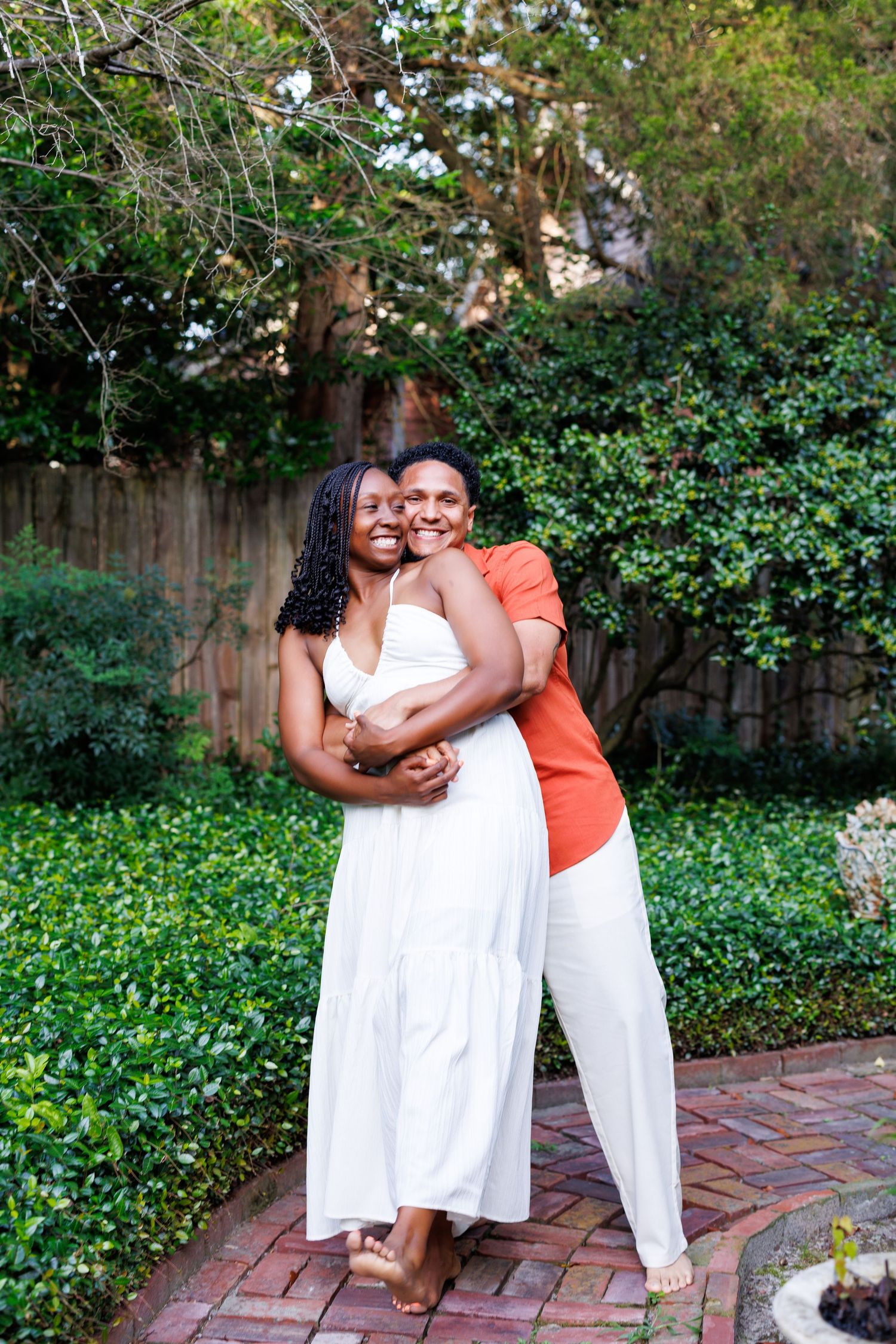 Two women embracing and smiling in a lush garden, one wearing white dress, other in orange top and white pants.