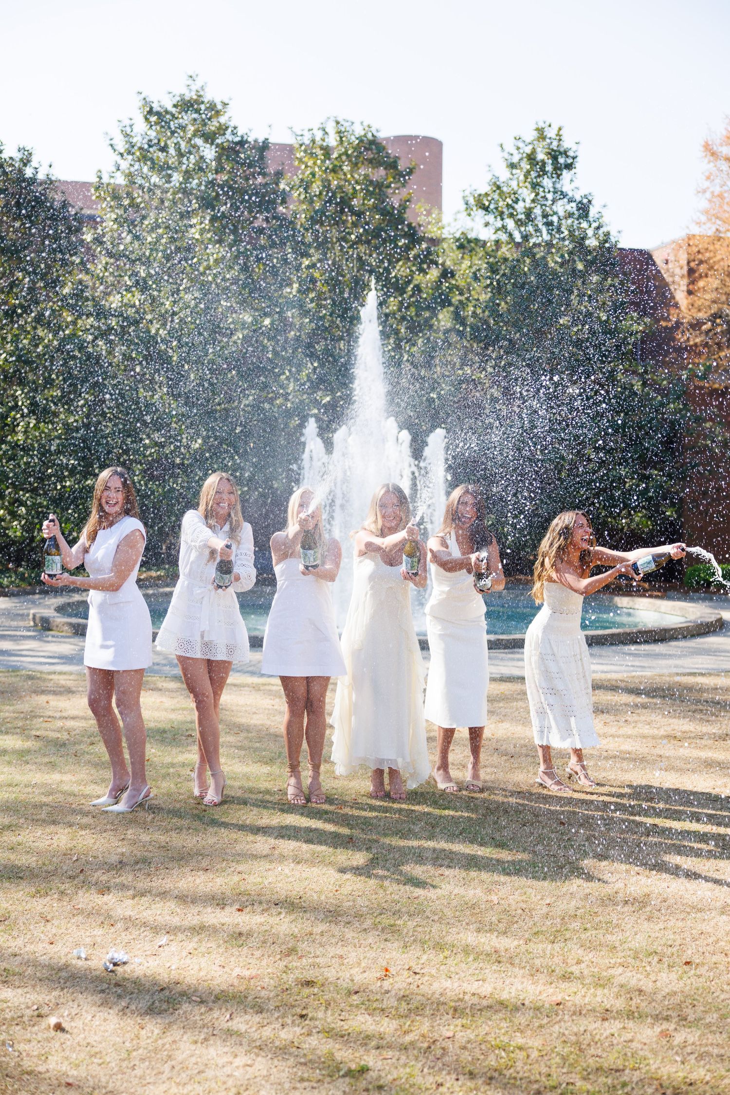 Bride and bridesmaids in white dresses celebrate with champagne by a fountain on a sunny day.