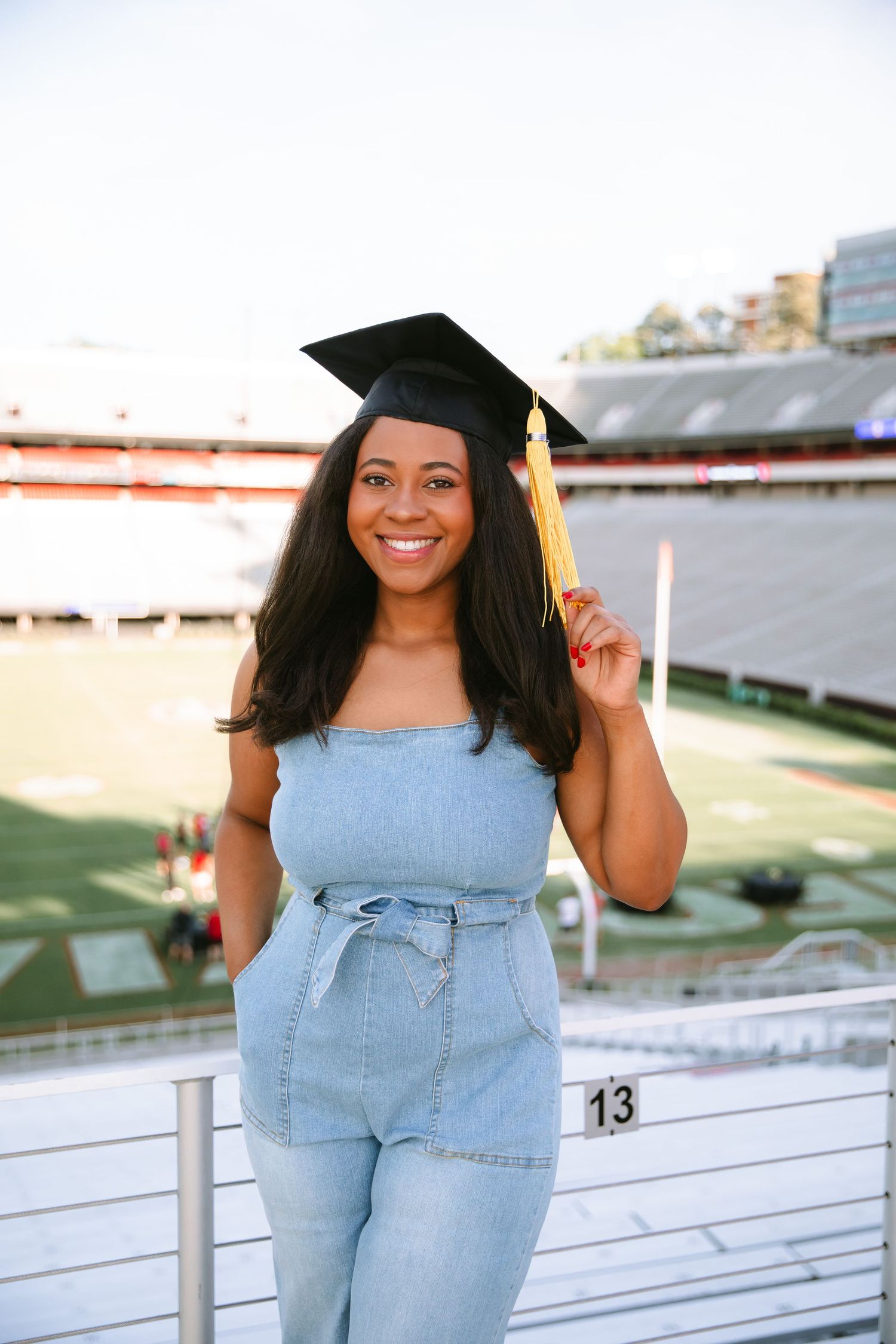 A graduate in a light blue denim outfit poses on stadium bleachers for graduation photos.