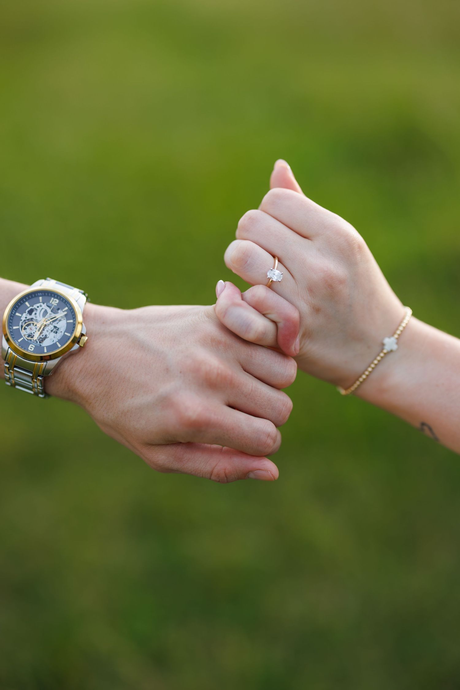 Couple holding hands showing engagement ring and luxury watch against green background.
