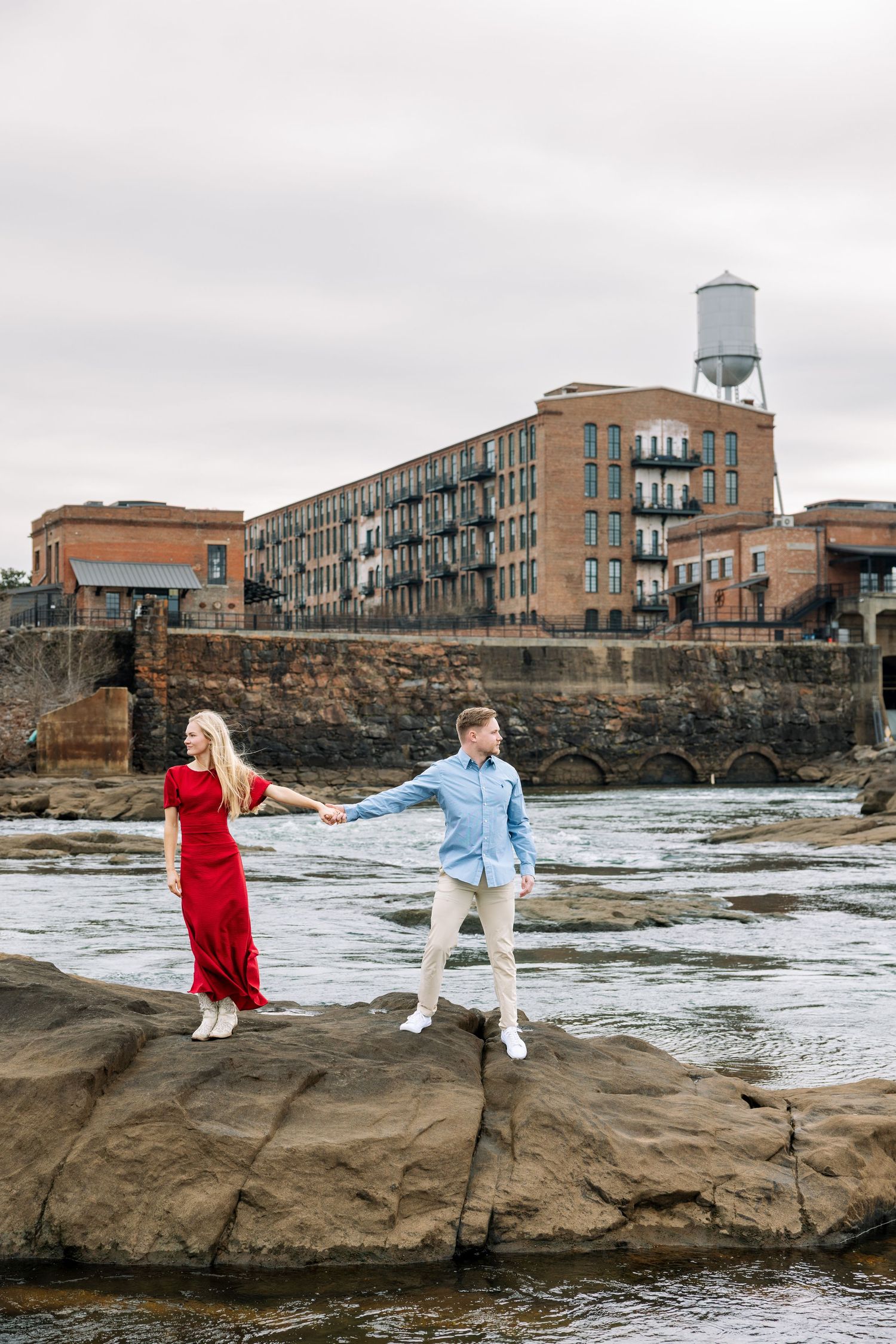 Playful couple poses on rocky shore with urban waterfront buildings in background.