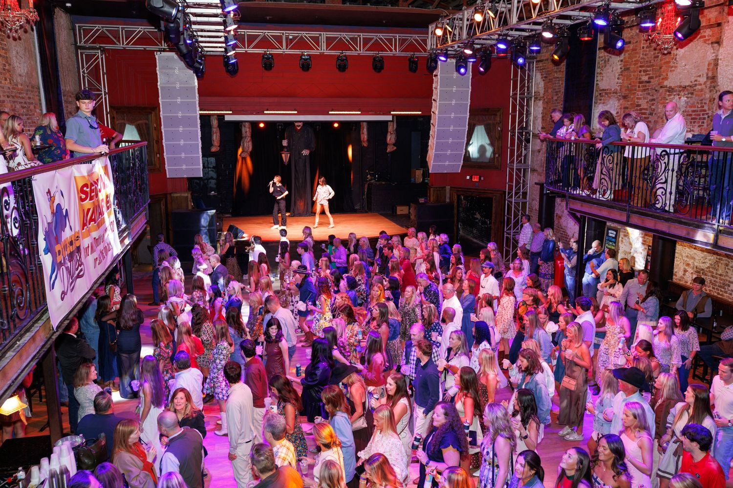 Packed nightclub dance floor with colorful stage lighting, live performer on stage, and crowd of people enjoying the event.