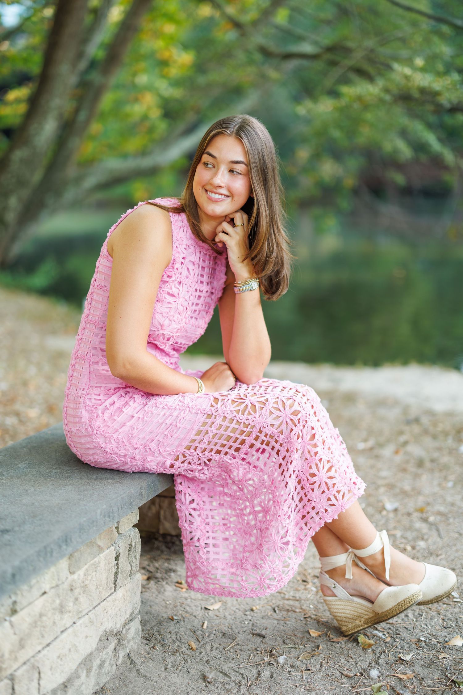 A person in a pink lace dress sits on a stone wall near a lake in a natural outdoor setting.
