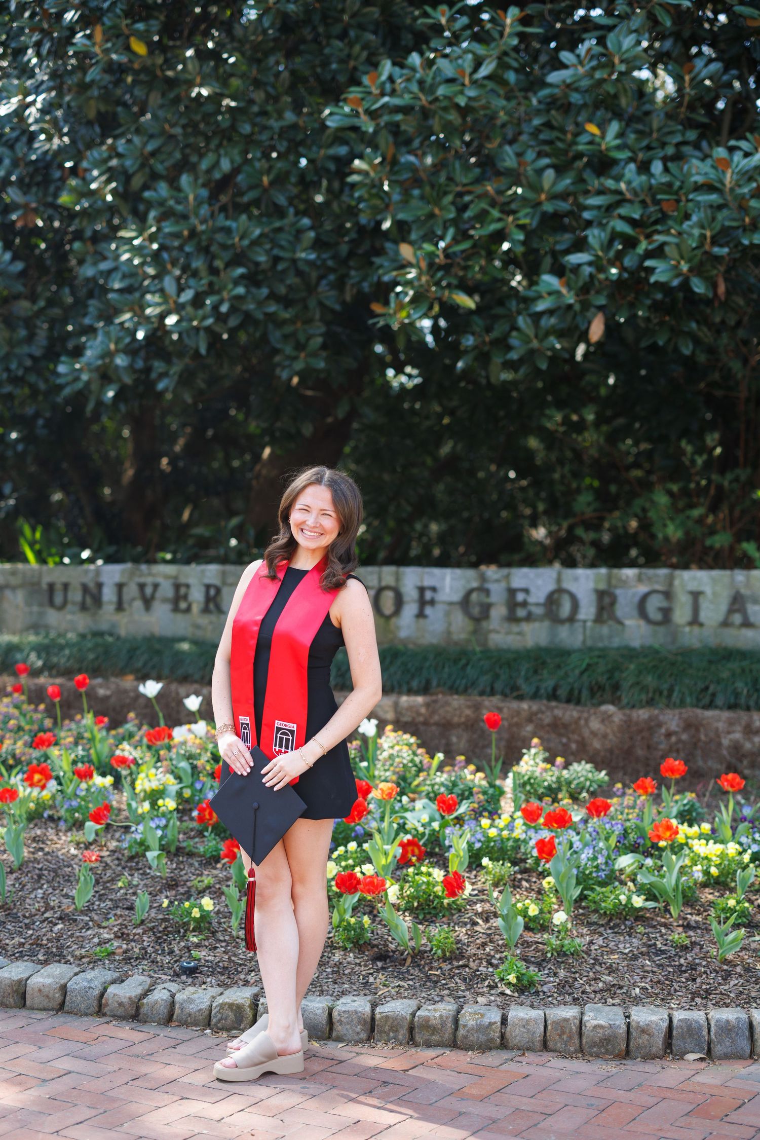 University of Georgia graduate smiling in black dress with red stole, posing by floral garden and UGA stone sign.