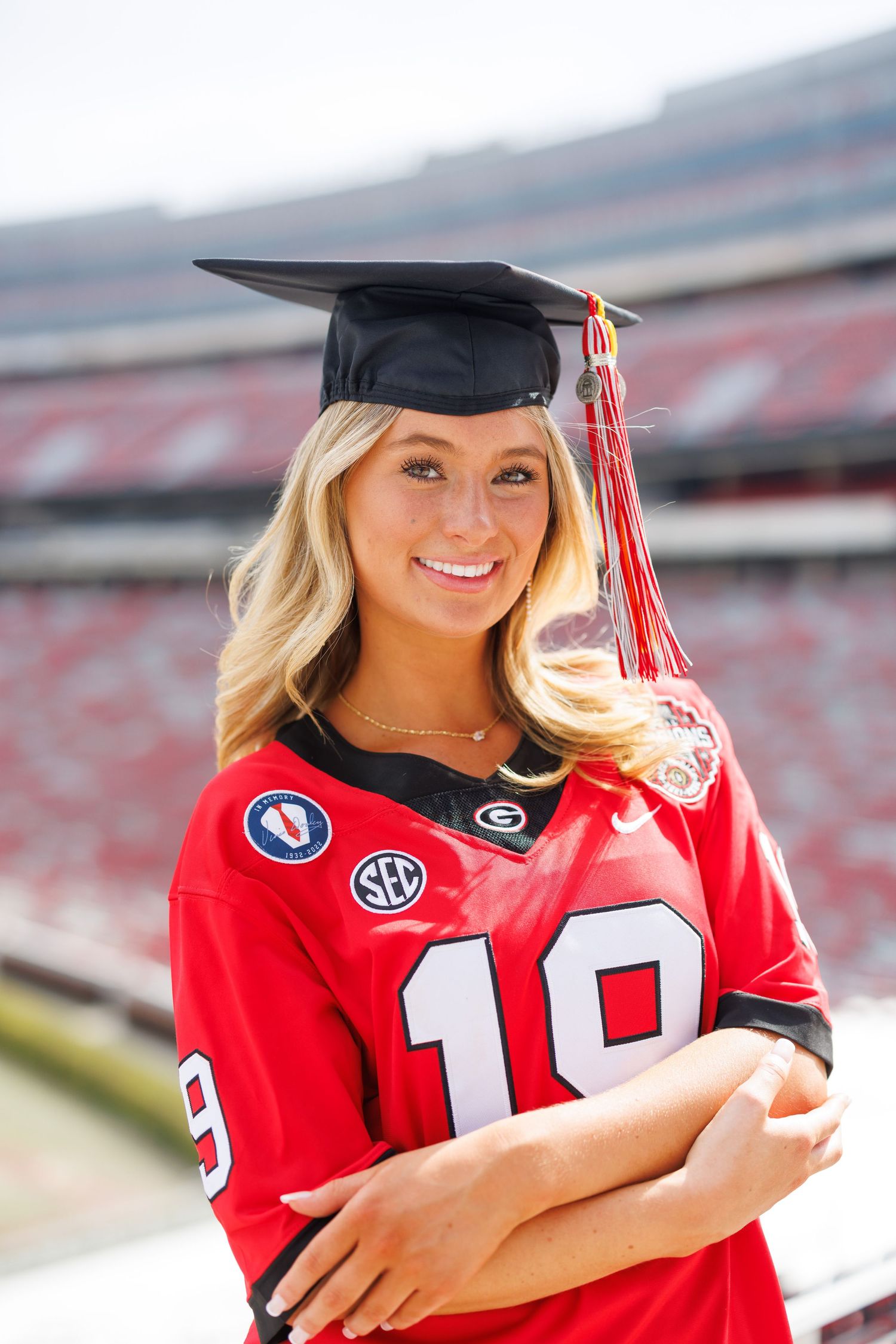 Graduate in red football jersey and graduation cap poses at stadium with tassel and bright smile.