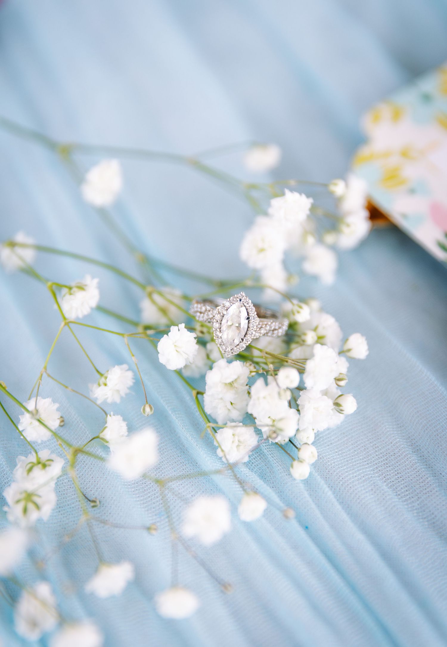 A delicate engagement ring nestled among white baby's breath flowers on blue fabric.
