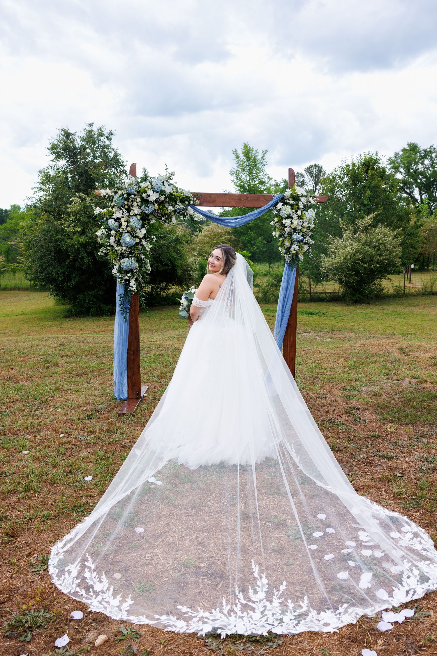 Bride in white gown with long lace veil stands before floral arch with blue ribbons at outdoor wedding ceremony.