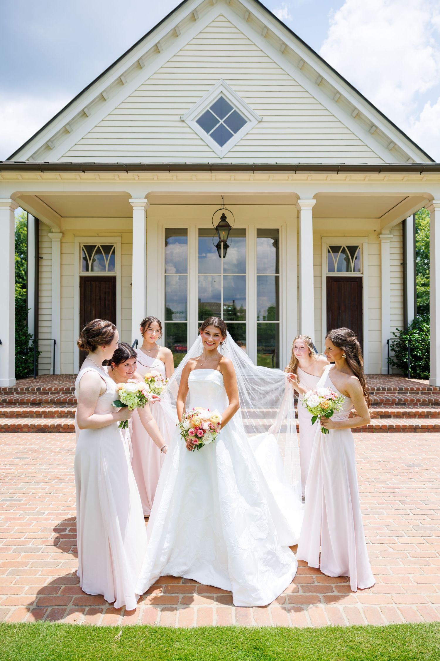 Bride in white gown and veil stands with four bridesmaids in blush dresses holding bouquets outside a white chapel.