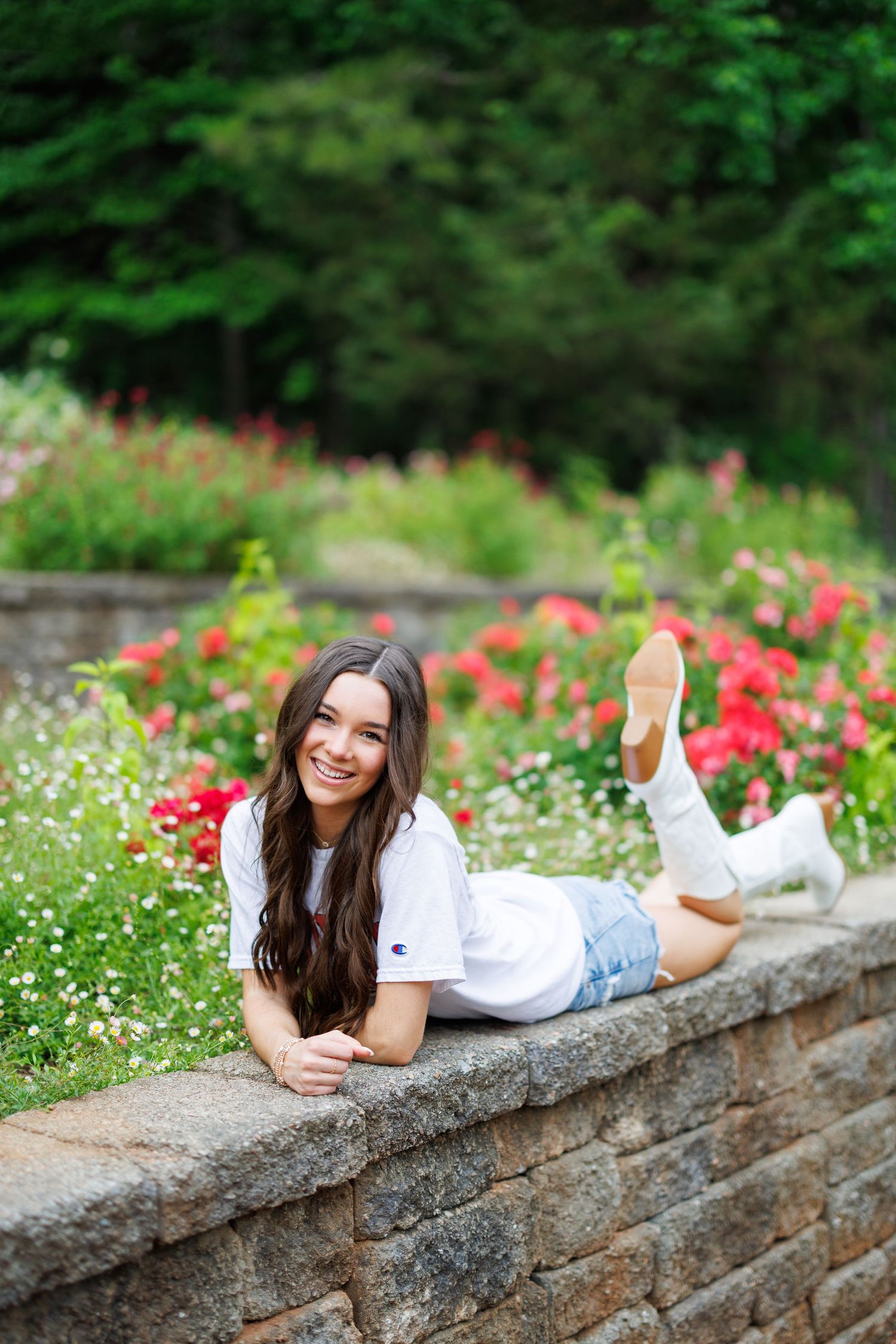 Young woman in white Champion tee and denim shorts posing on stone wall surrounded by blooming red flowers.