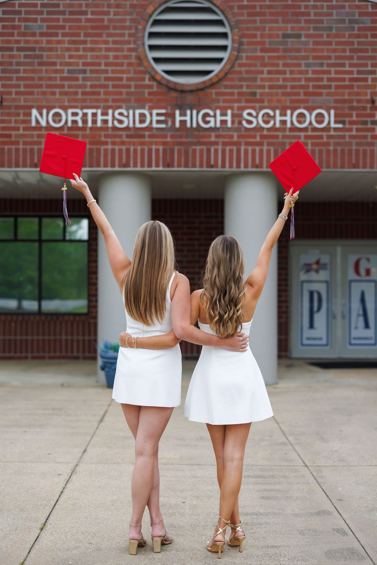 Two female graduates in white dresses raise red caps in front of Northside High School brick building.