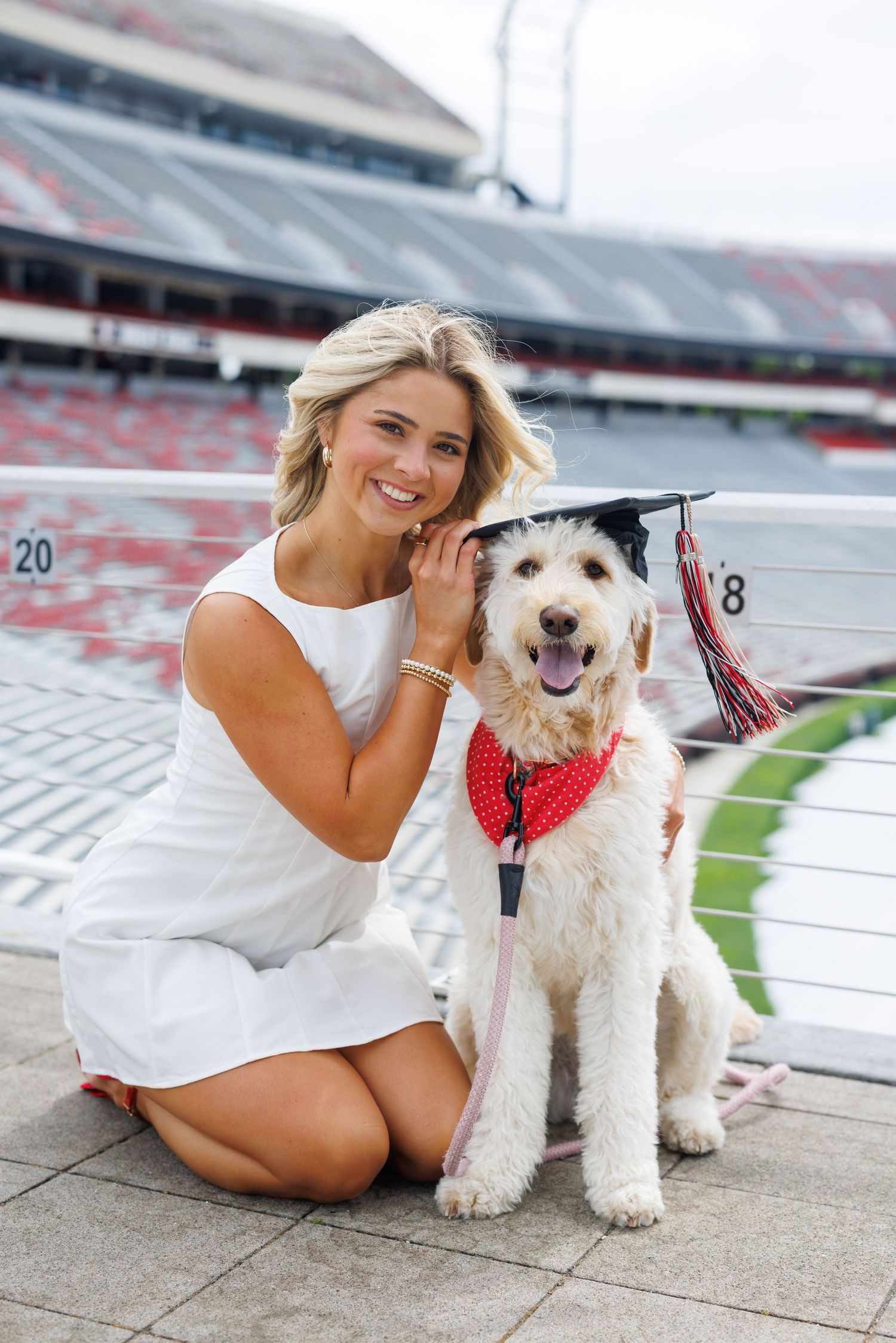 Smiling blonde woman in white dress poses with a Goldendoodle wearing a graduation cap and red bandana in a stadium.