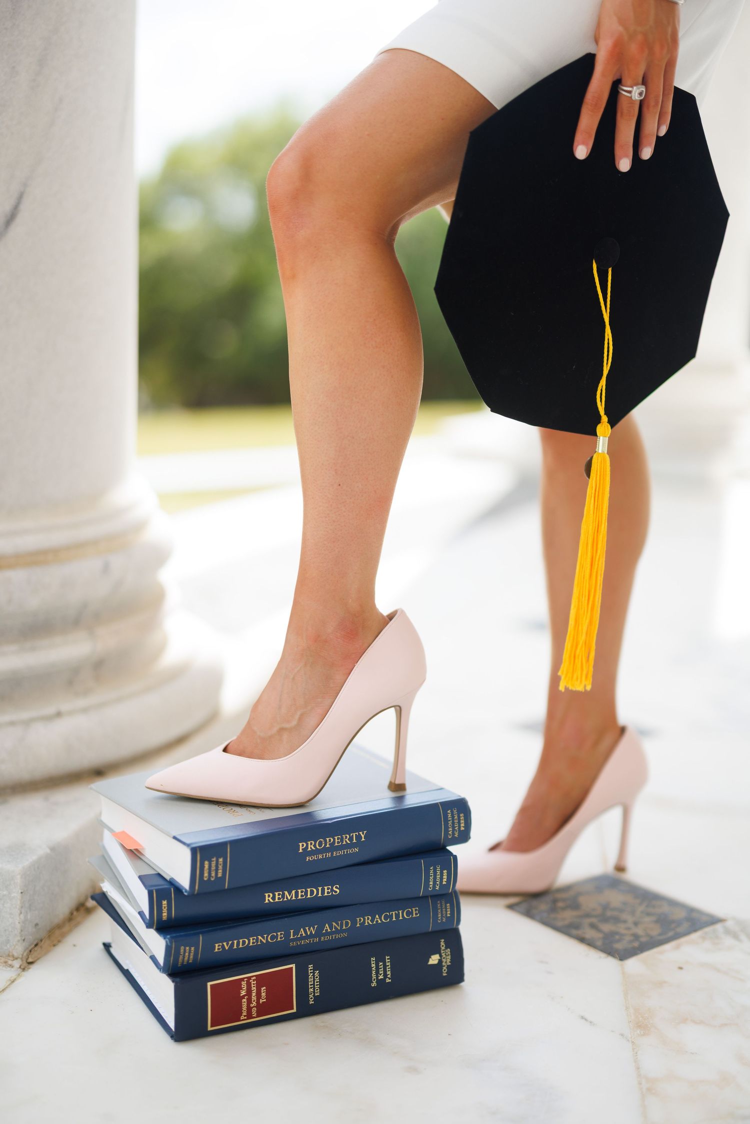 Law school graduate in pink heels stands on stacked legal textbooks holding black mortarboard cap with gold tassel.