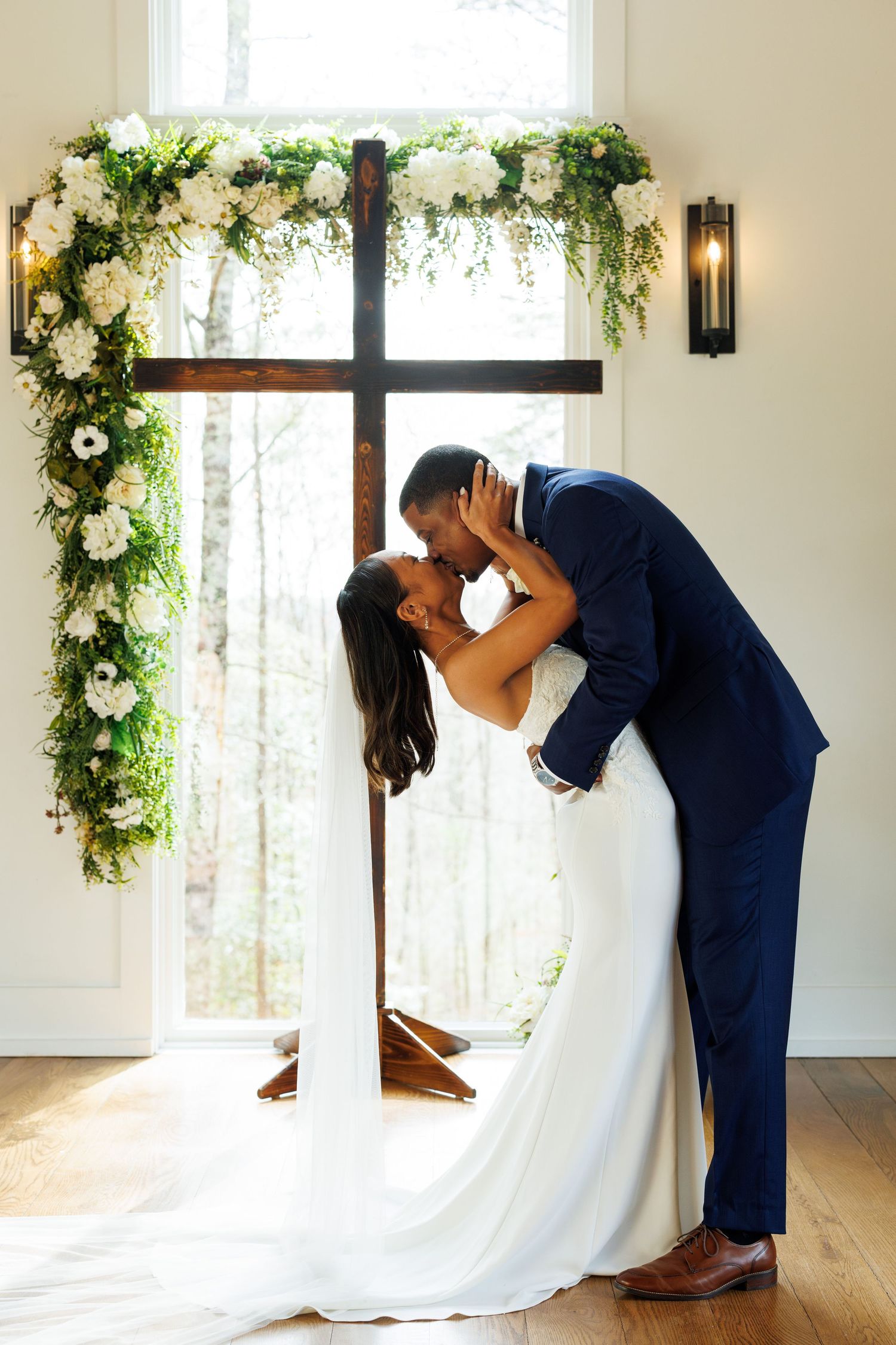 A romantic wedding kiss under a floral arch with white flowers and greenery in an elegant indoor setting.