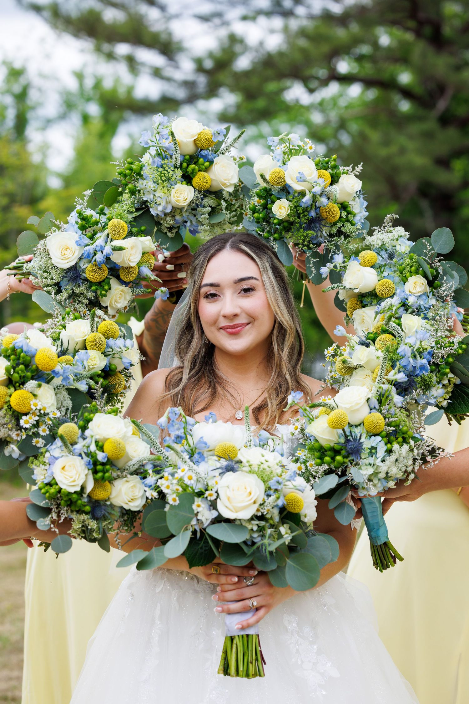 Bride smiling through a floral frame of blue, white, and yellow bouquets held by bridesmaids in yellow dresses.
