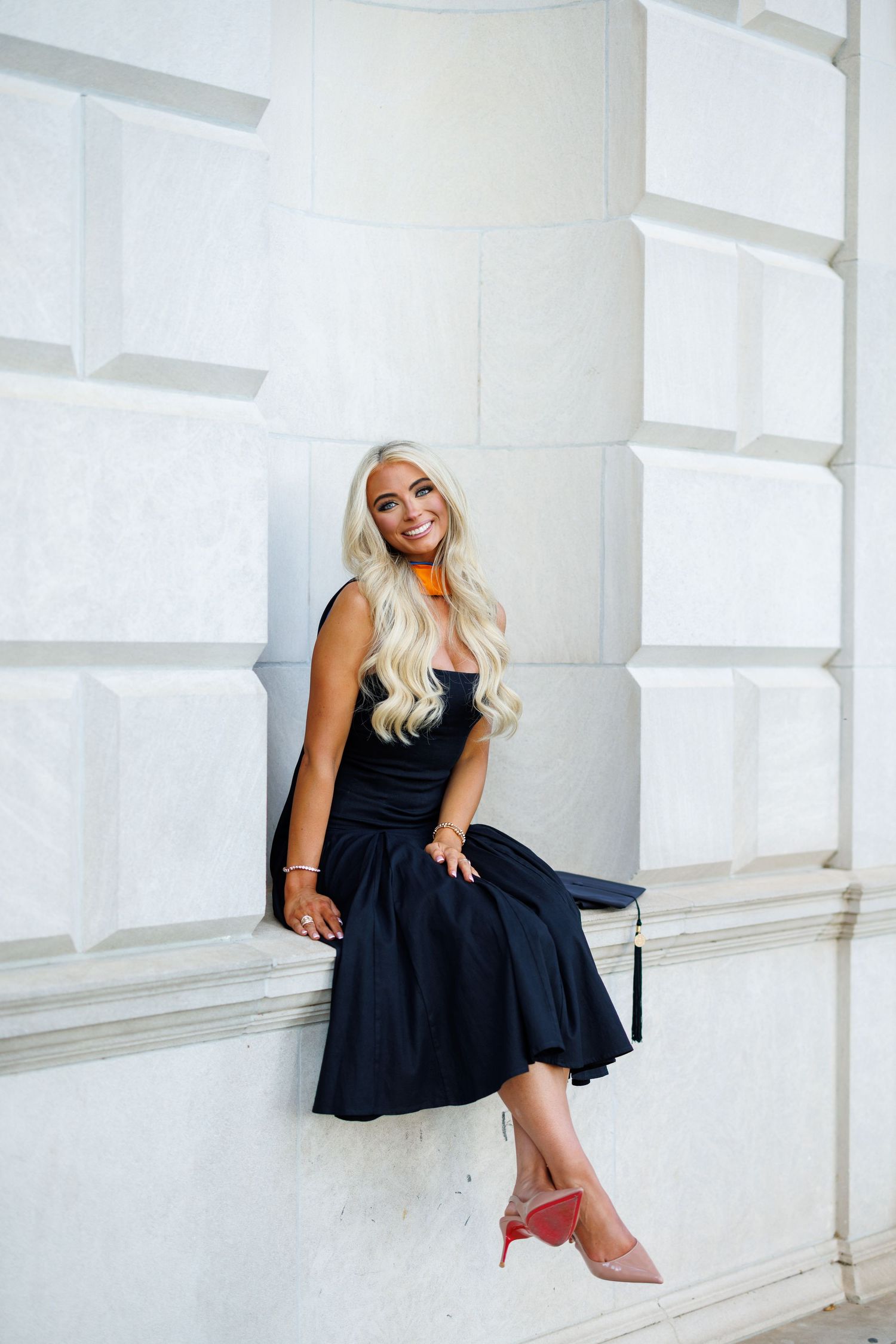 Smiling blonde woman in black dress holding graduation cap, seated against white architectural wall.