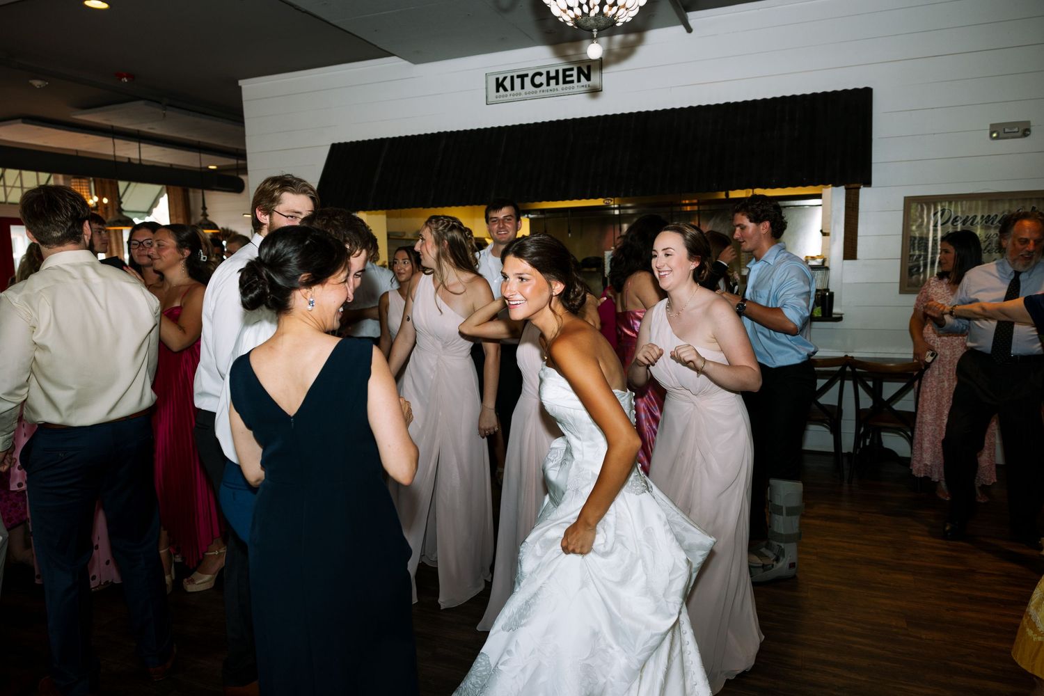 Bride in white gown dancing with bridesmaids in pink dresses at a lively wedding reception near the kitchen.