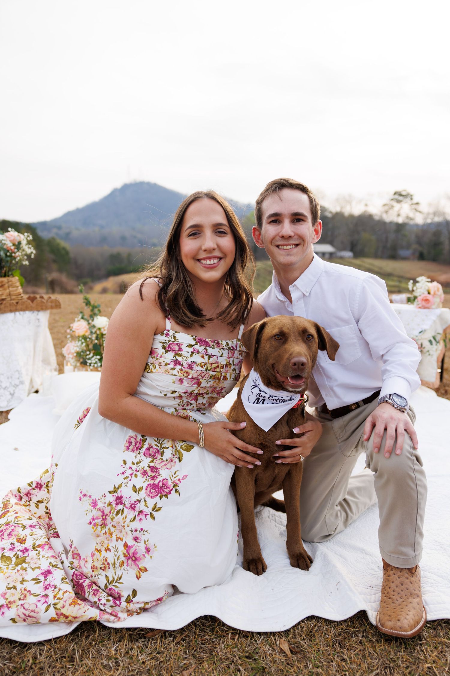 A couple sits outdoors with their brown dog wearing a bandana against a mountain backdrop.