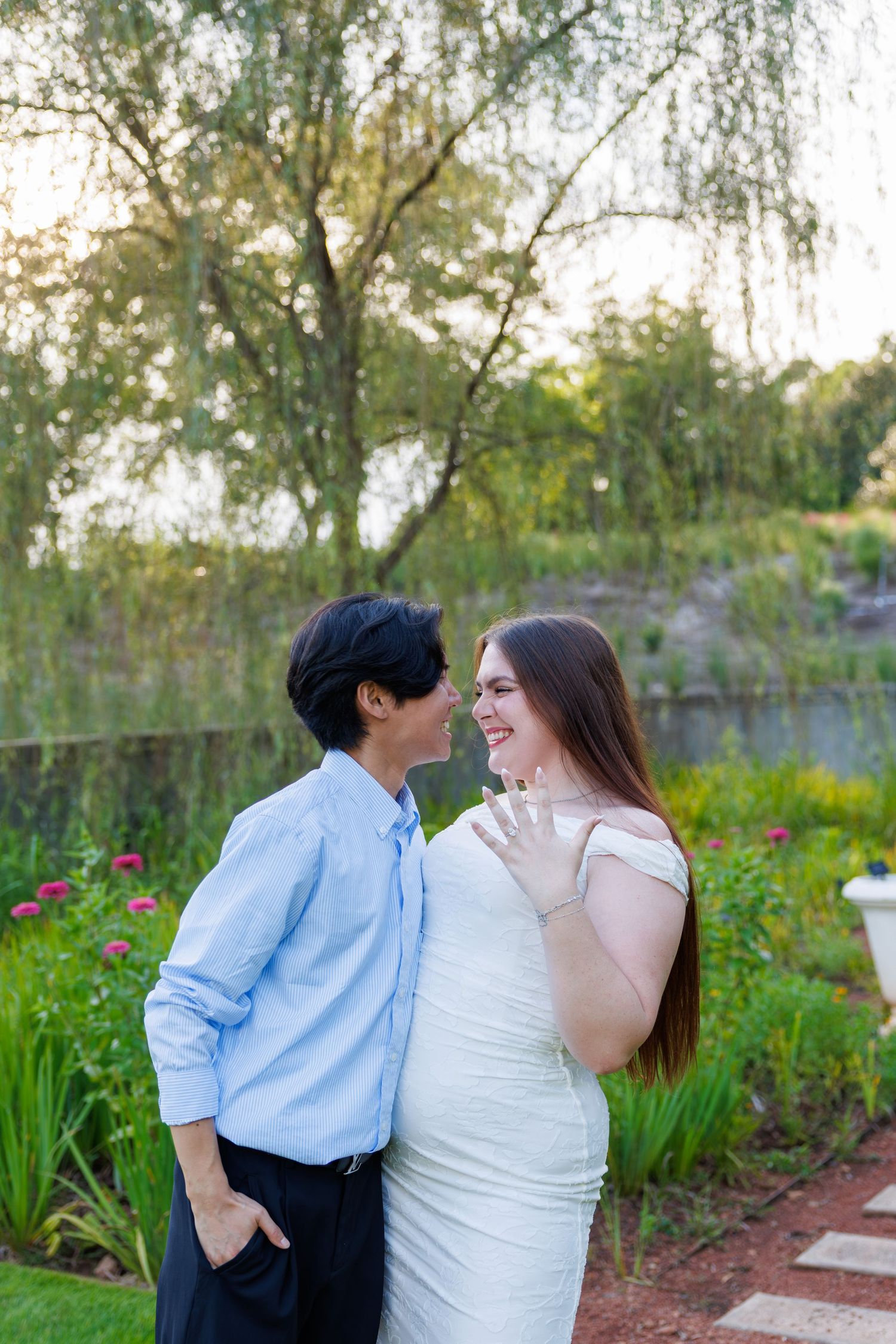 Couple in love smiling at each other outdoors, woman in white dress showing engagement ring, lush garden and pond backdrop.