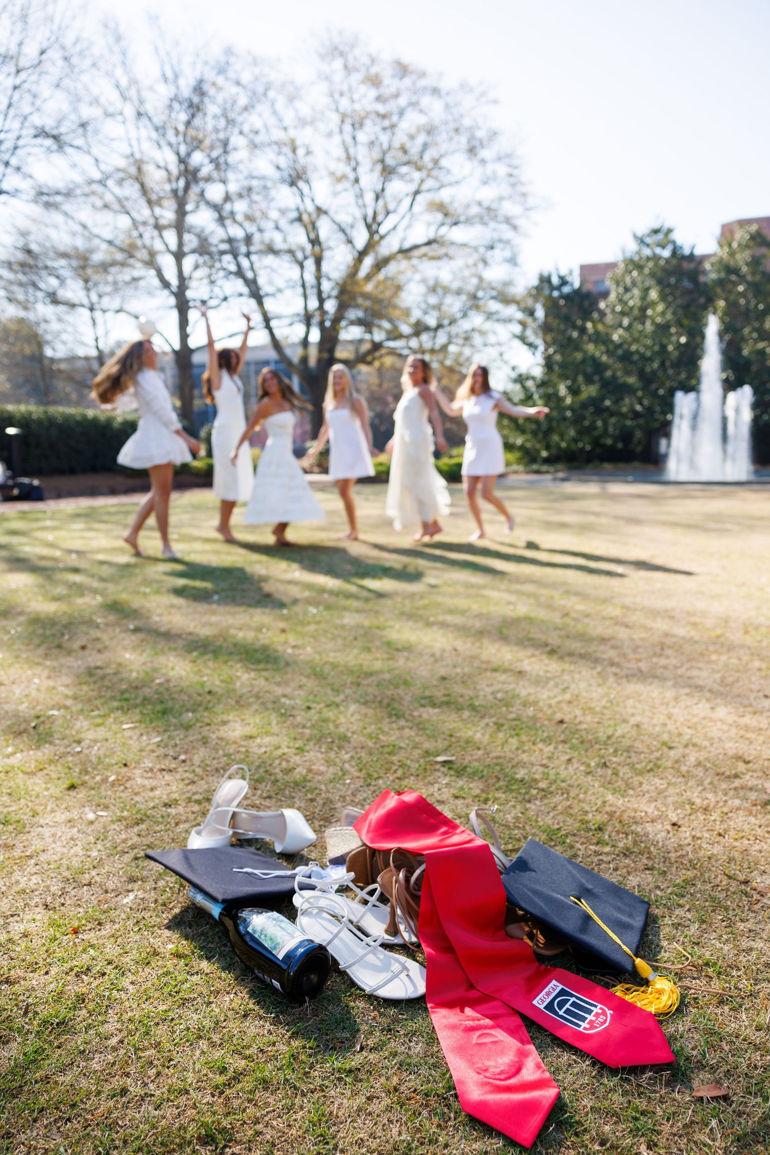 People in white dresses running and jumping in a field while their shoes and accessories lay scattered on the grass.