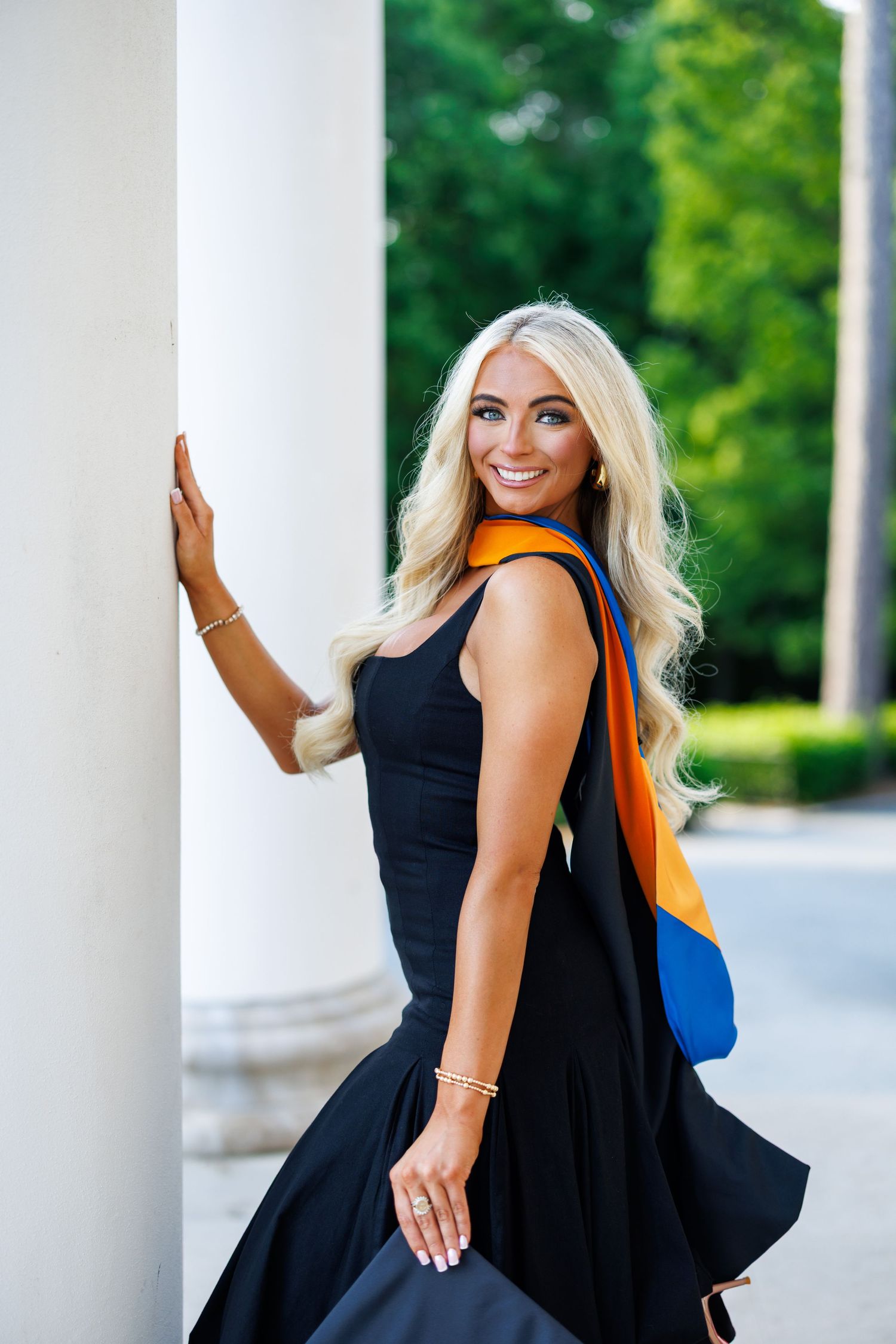 Smiling blonde woman in black dress with blue and orange graduation stole leaning against white column outdoors.