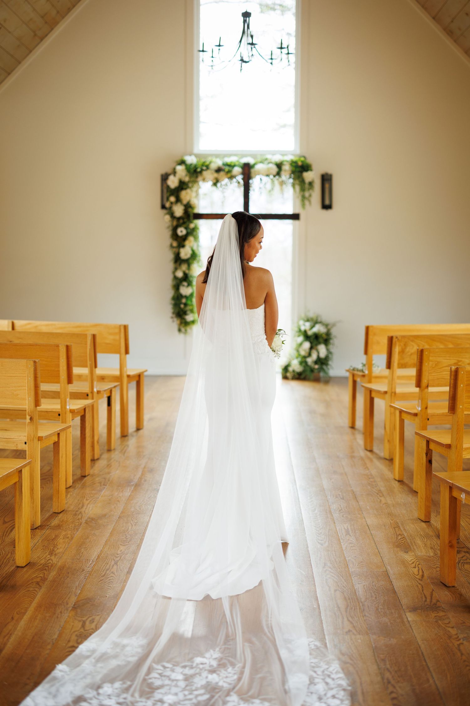 A flowing white wedding dress and veil captured from behind in a rustic chapel with wooden pews.