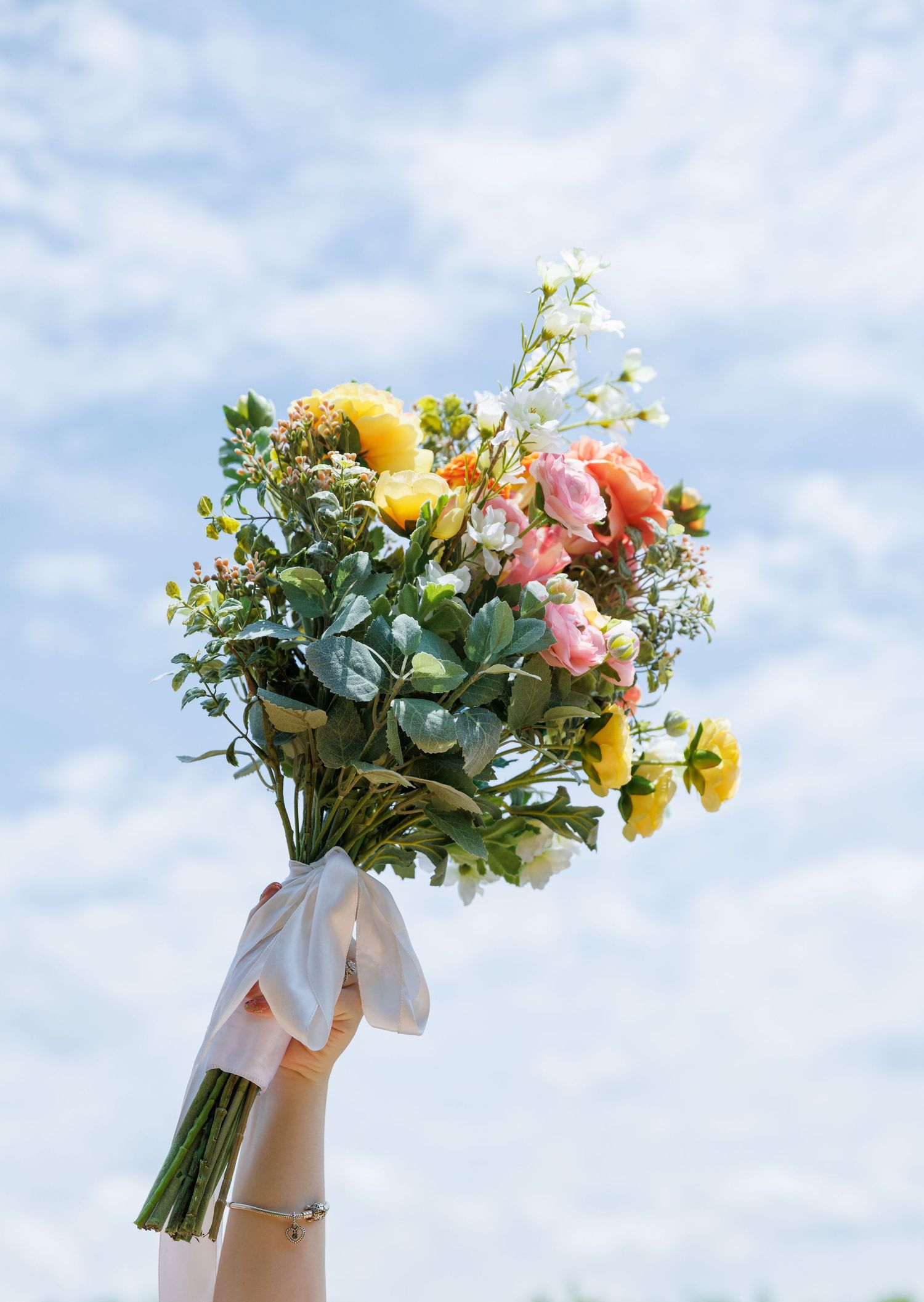 A vibrant spring wedding bouquet with yellow and pink flowers held up against a blue sky.