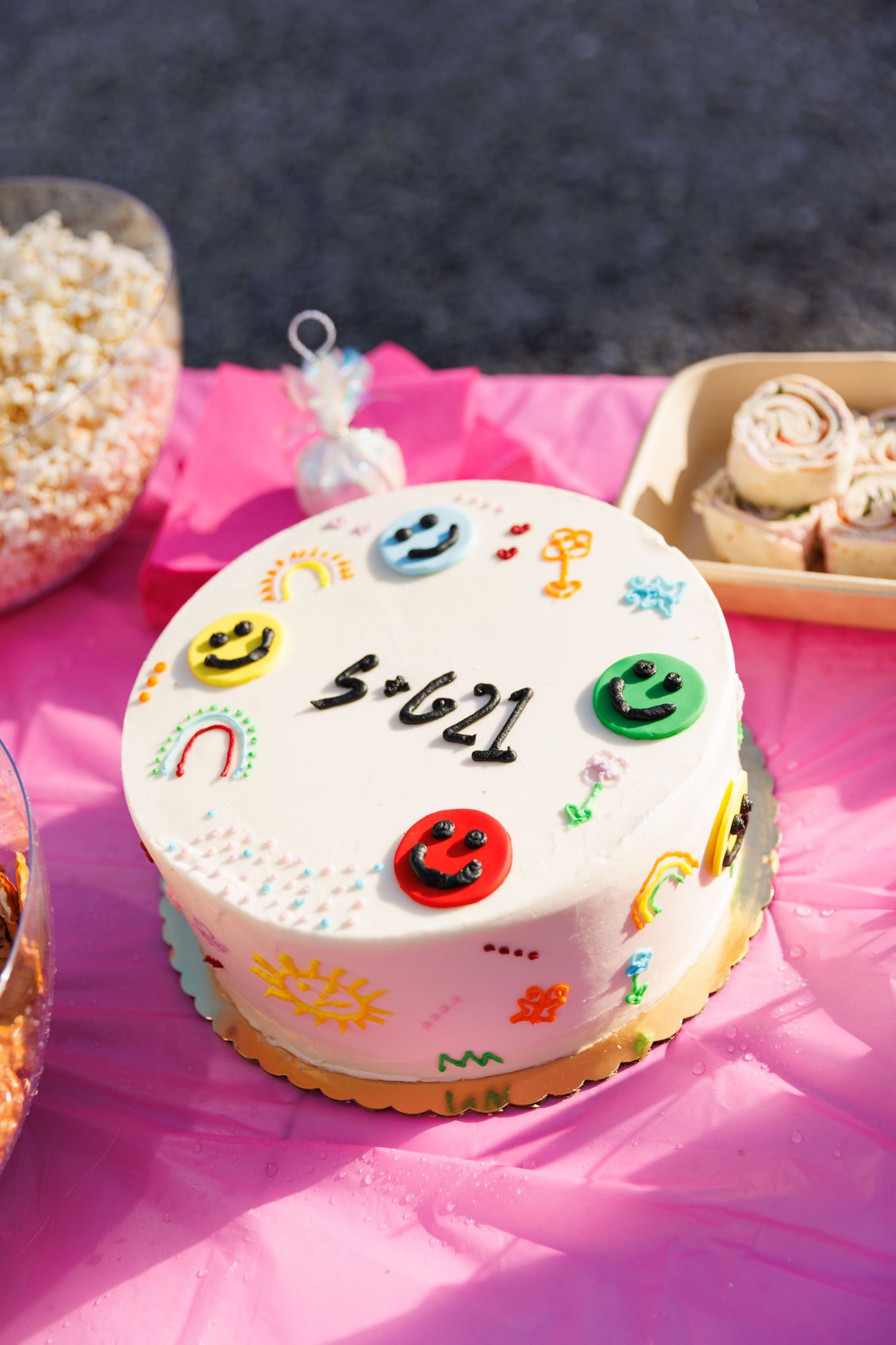 White birthday cake with smiley face decorations and colorful designs on a pink tablecloth.