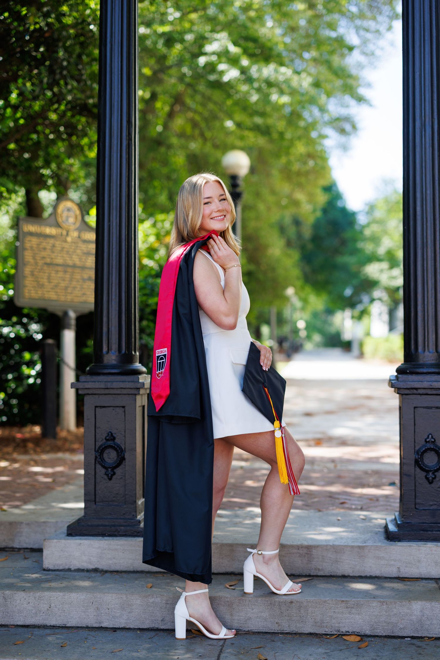 Smiling female graduate in white dress holding black cap and gown with red sash, posing between ornate columns outdoors.