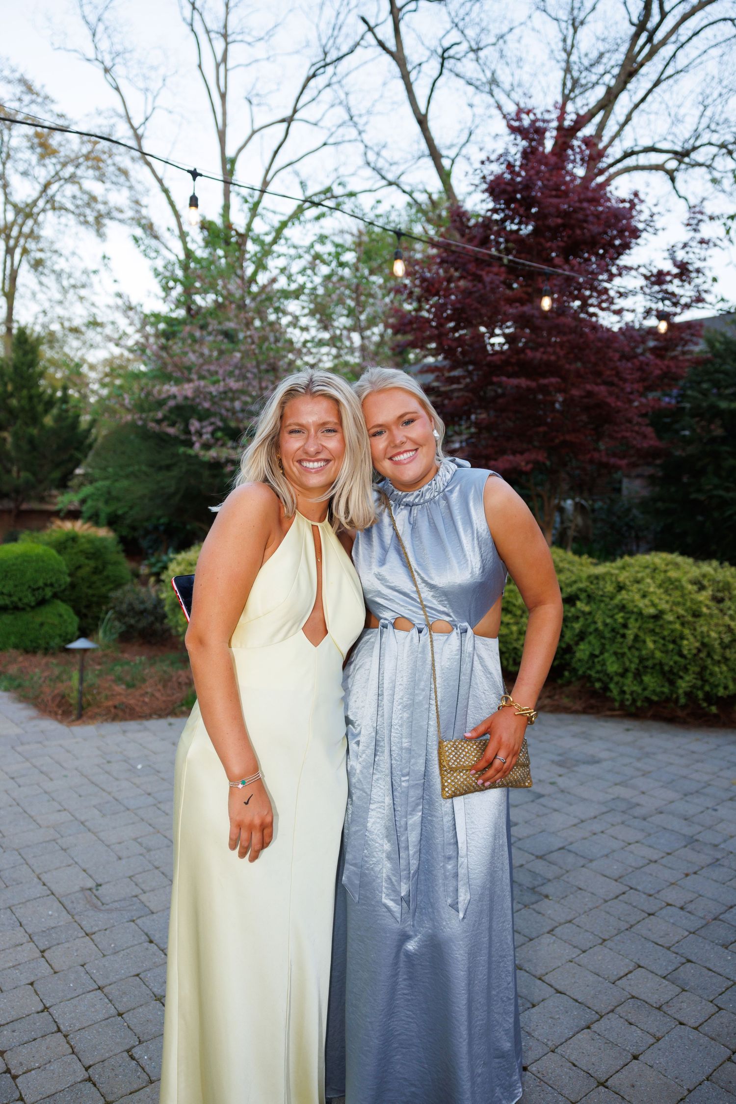 Two blonde women in elegant formal gowns pose together outdoors at a garden event.