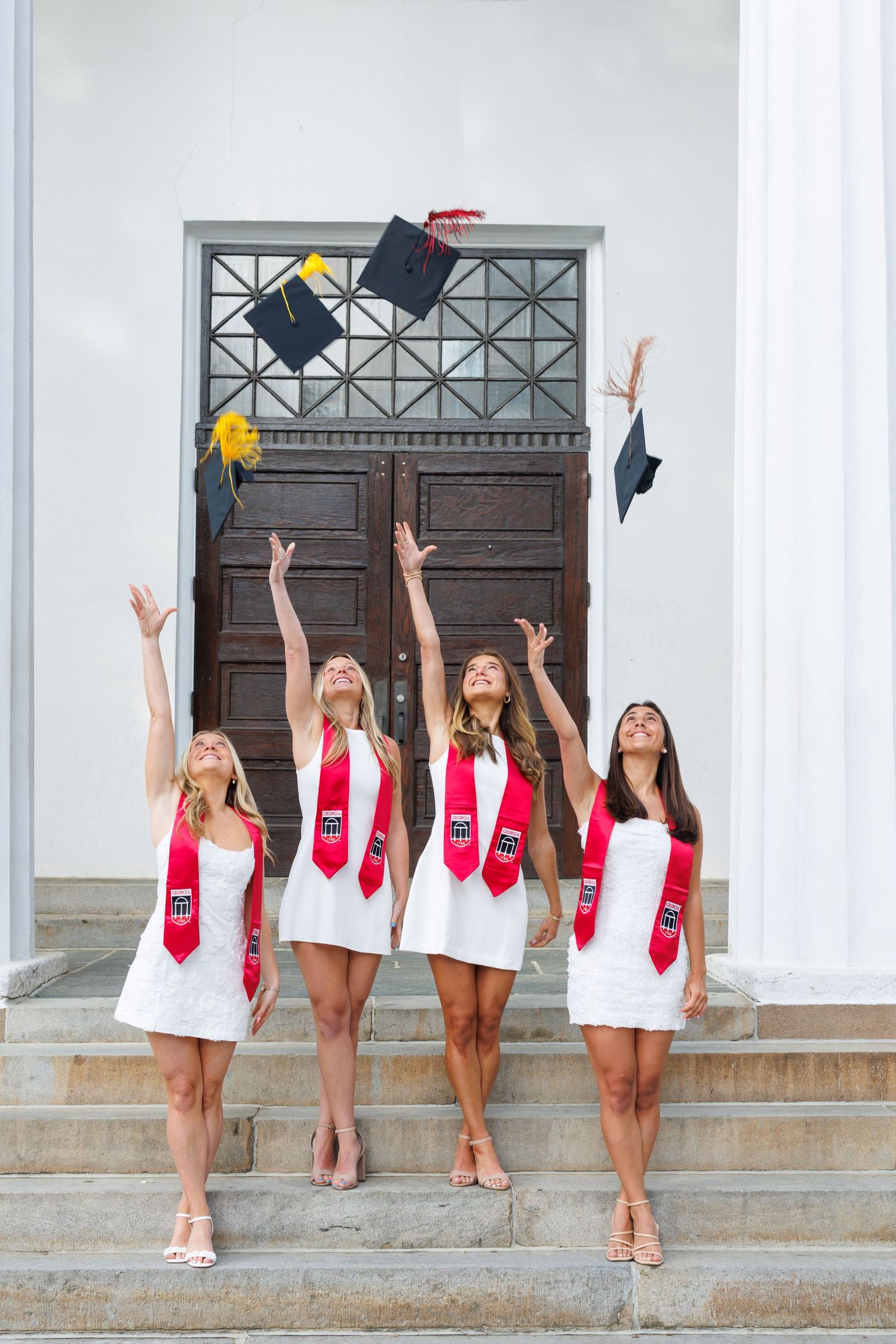 Four female graduates in white dresses and red stoles toss mortarboard caps on university steps.