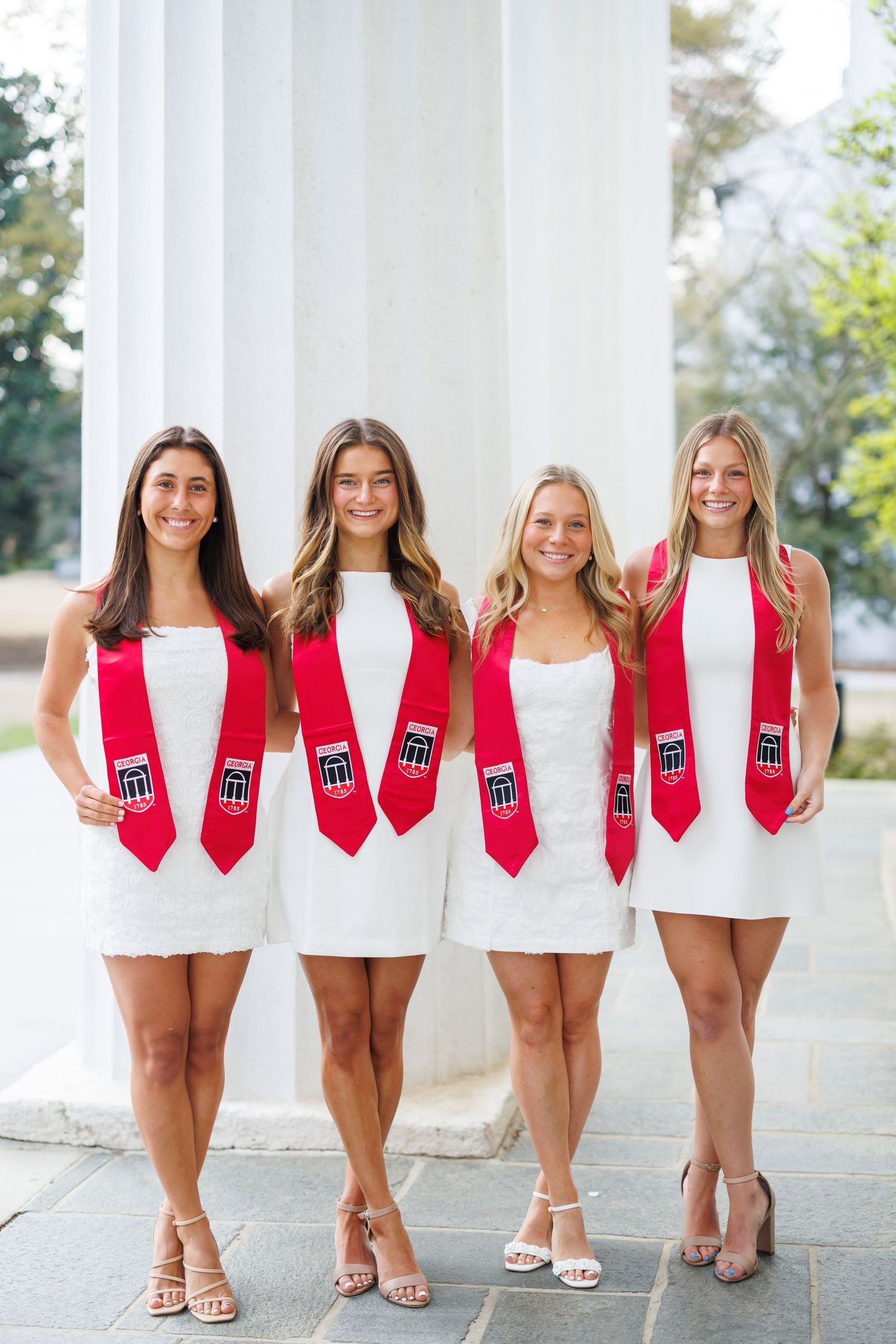 A group of sorority sisters wearing white dresses and red sashes pose together for graduation photos.