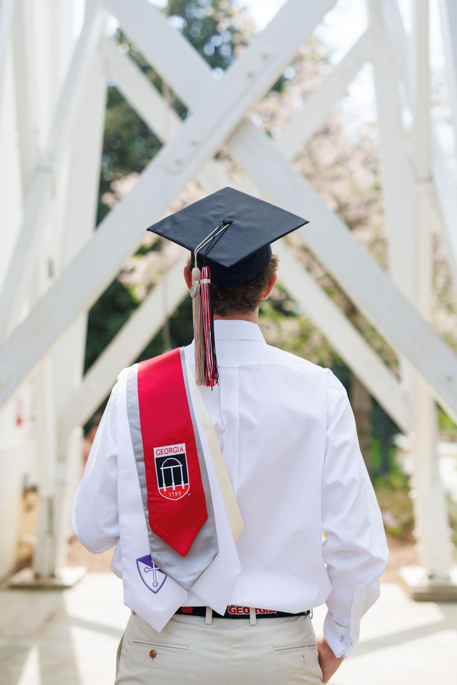 Graduate in white shirt wearing black mortarboard cap and red academic stole stands on bridge at commencement.