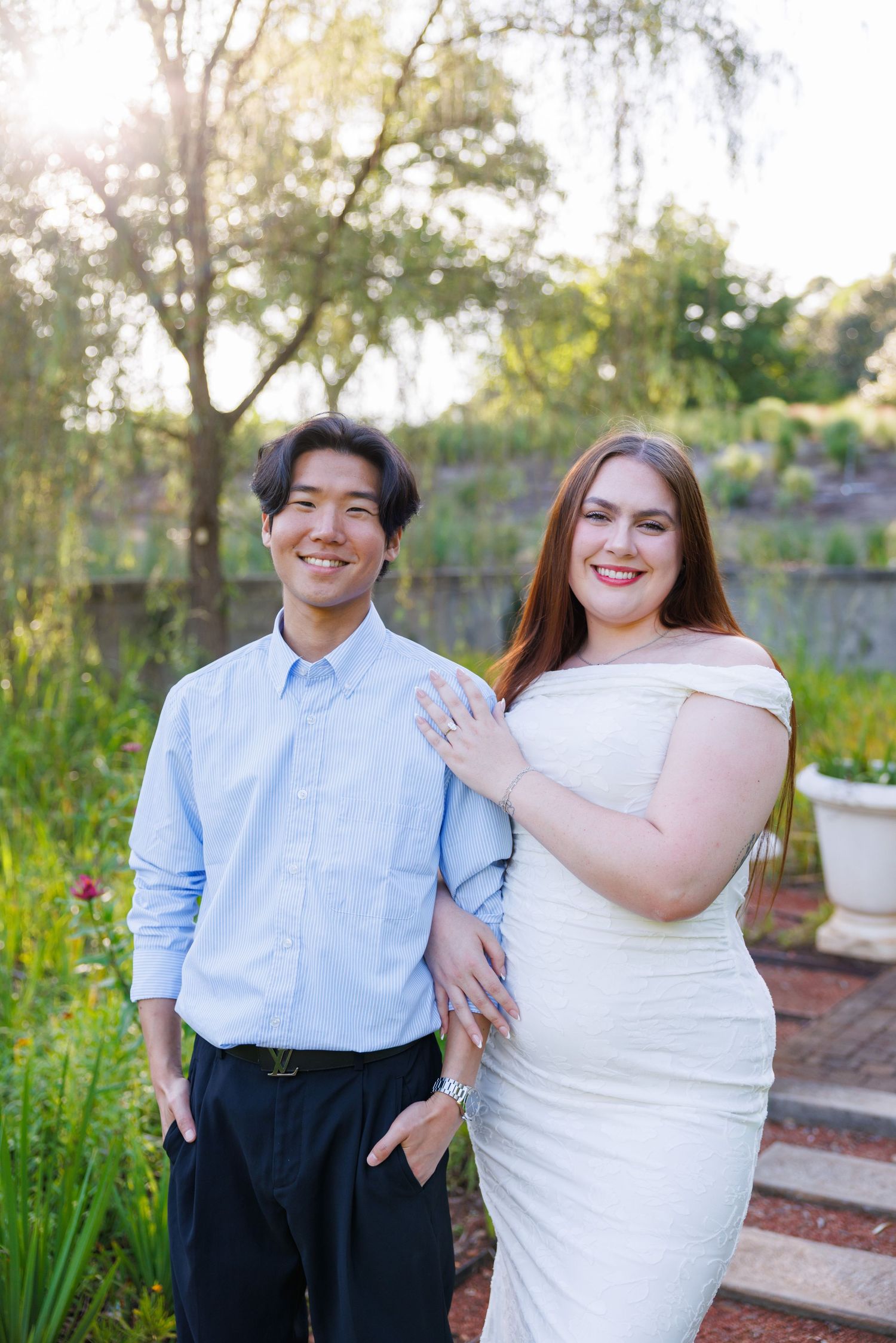 Smiling couple posing outdoors in garden, man in blue shirt, woman in white off-shoulder dress showing engagement ring.