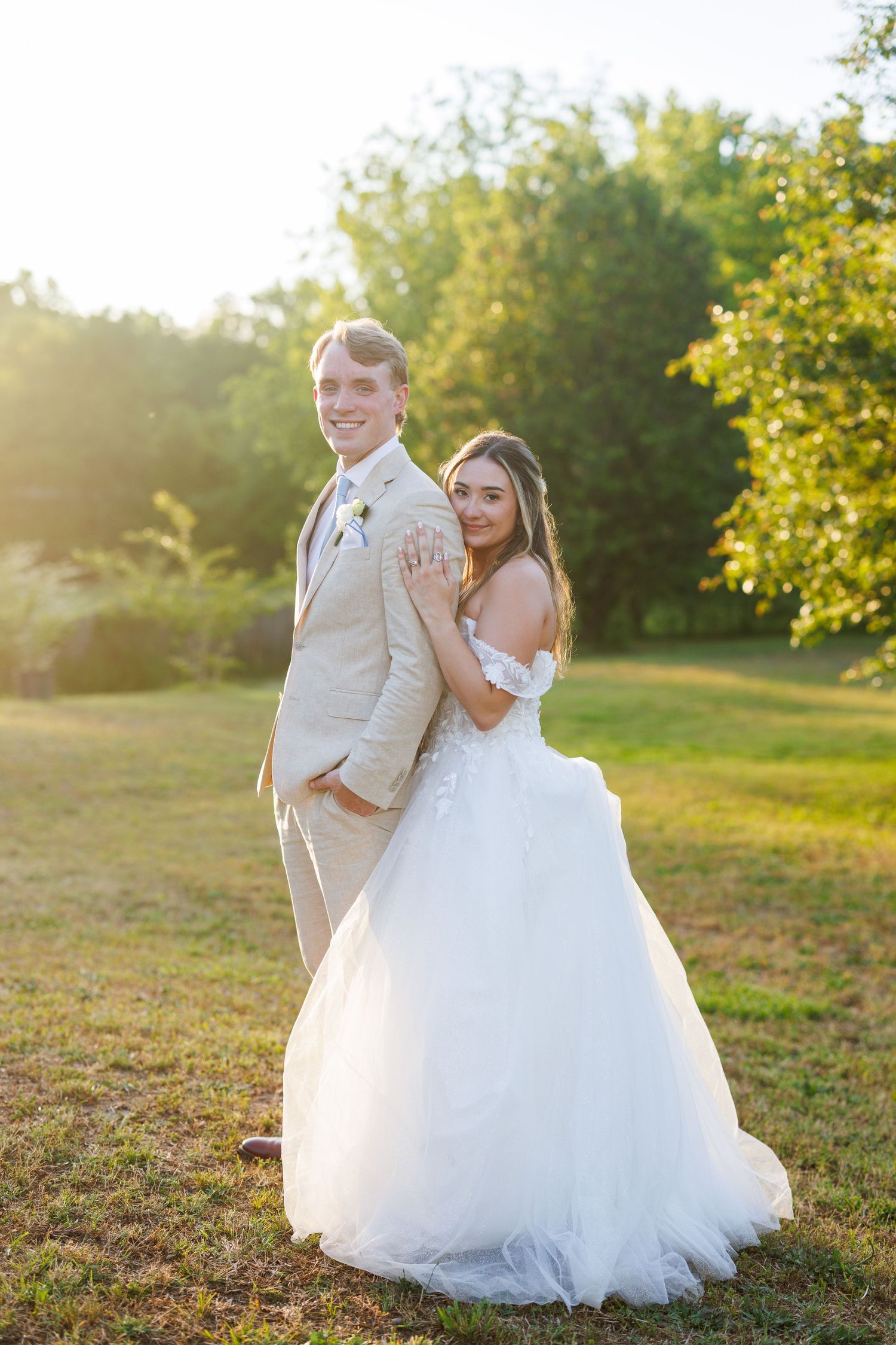 Bride in white gown and groom in tan suit pose back-to-back outdoors during golden hour sunset.
