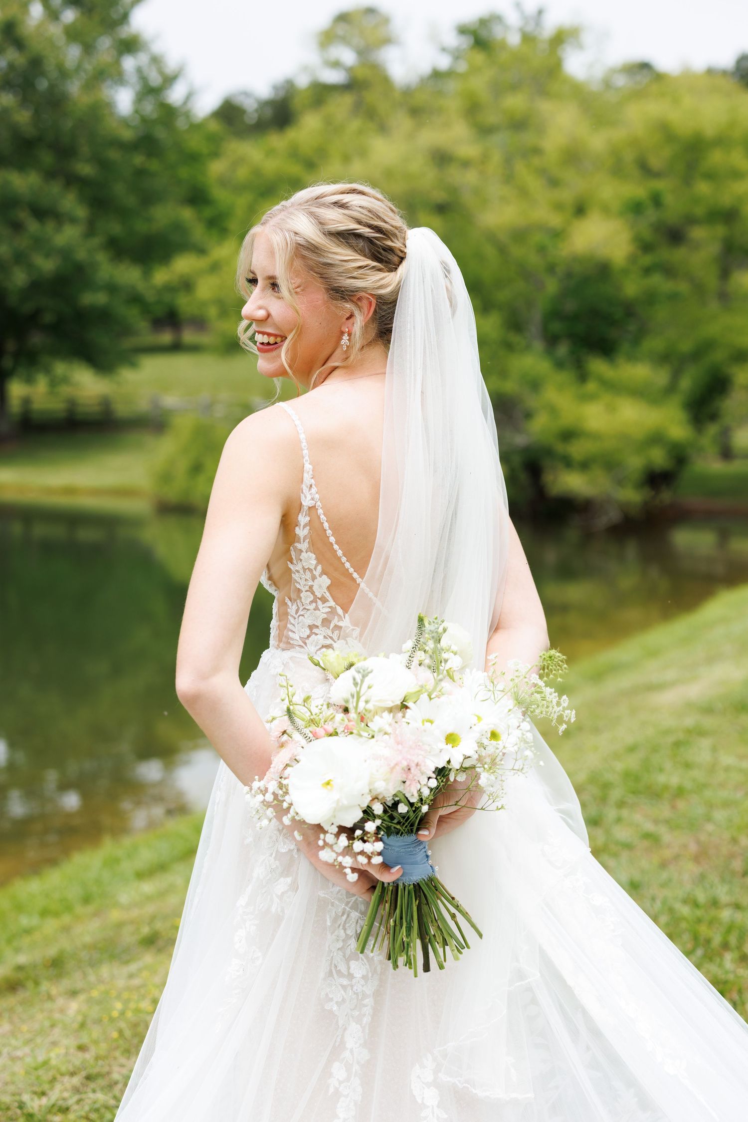 Bridal portraits showing the back details of a lace wedding gown with a bouquet against a lakeside backdrop.