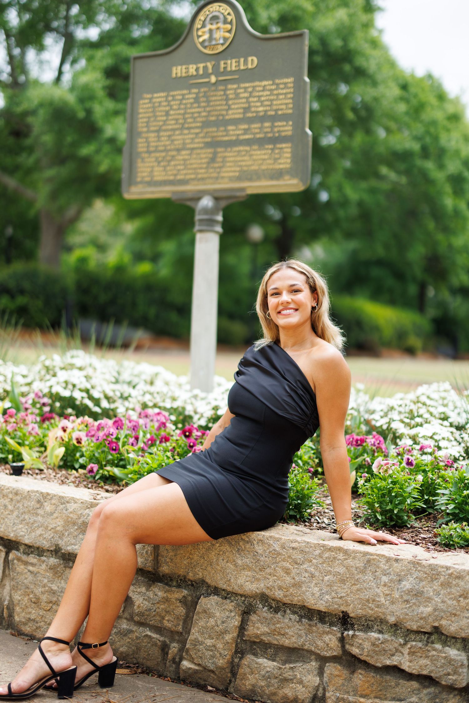 Graduate poses on stone wall near historical marker and garden flowers.