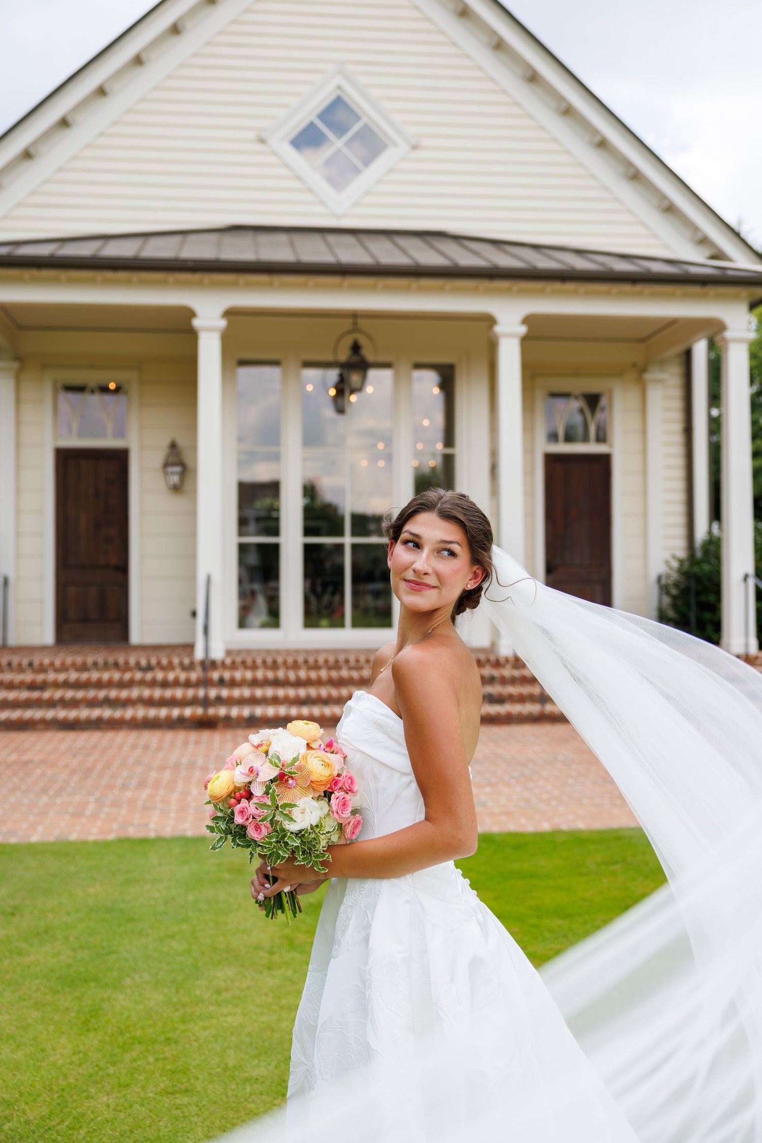 Bride in white strapless gown holds colorful bouquet, veil flowing, standing before elegant Southern venue.