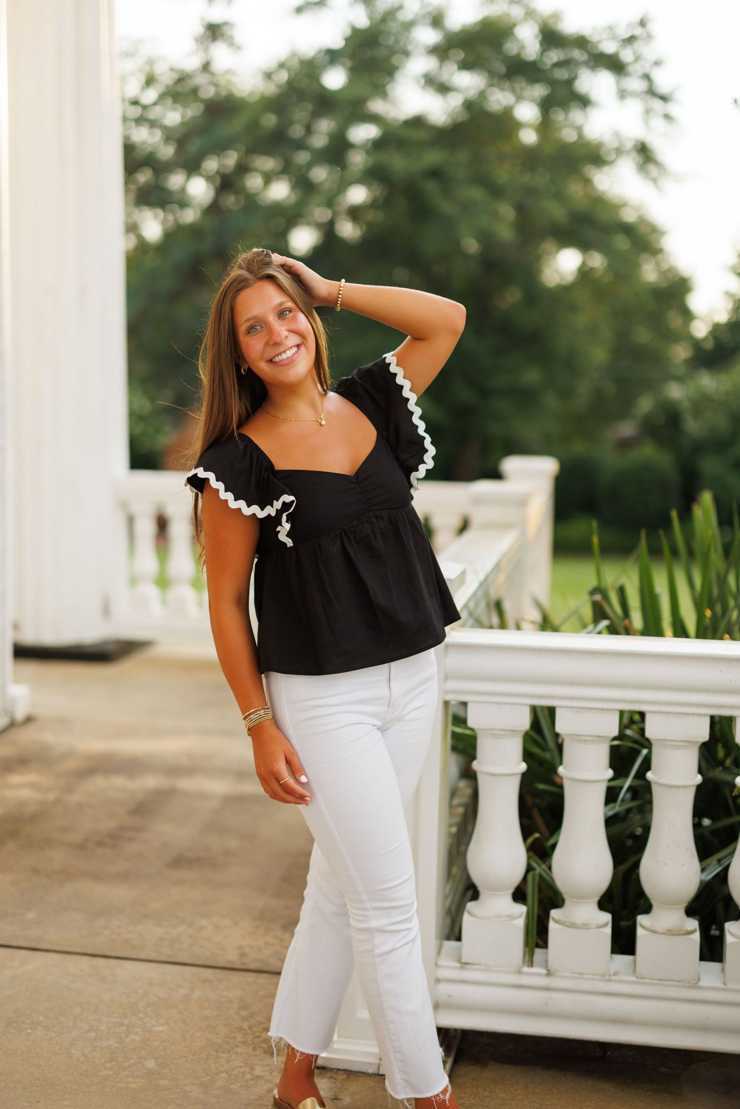 A woman in black and white attire poses casually on a porch with white columns and railings in the background.