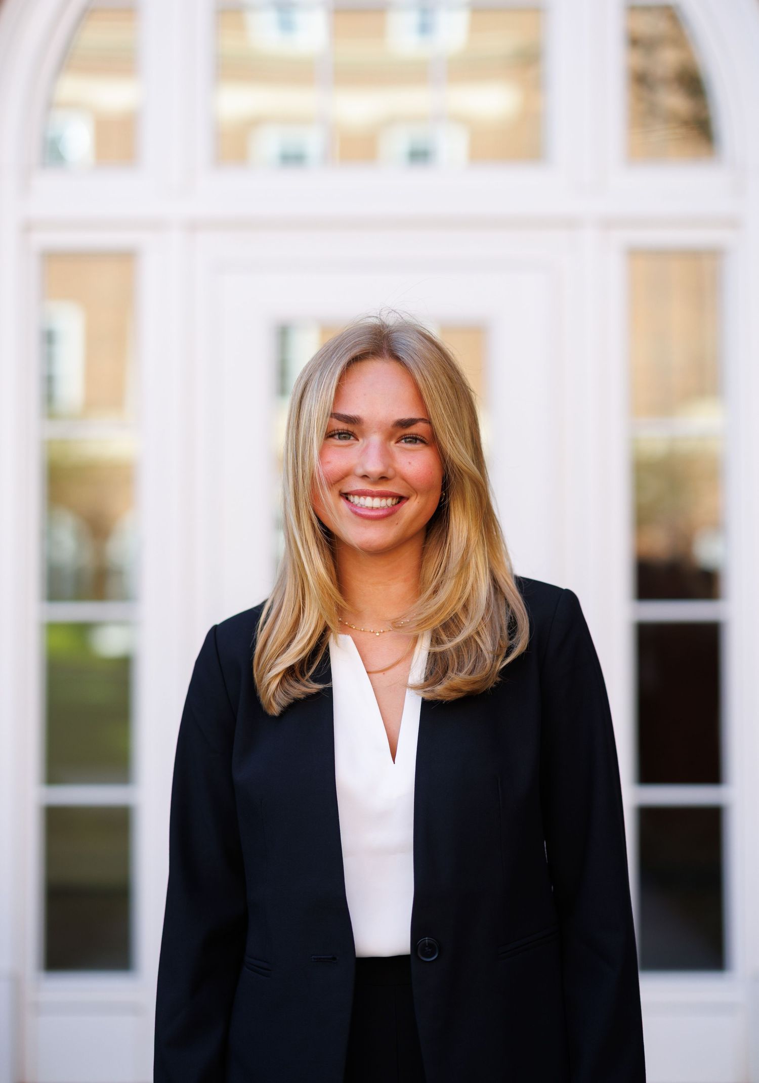Young blonde woman in black blazer and white top smiling in front of elegant white building entrance.