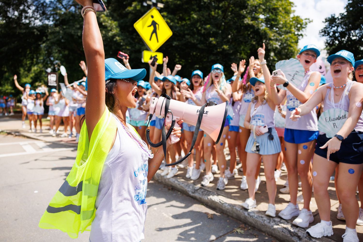 A crowd of enthusiastic students in blue caps cheers as a woman in a safety vest raises her hand during an outdoor event.