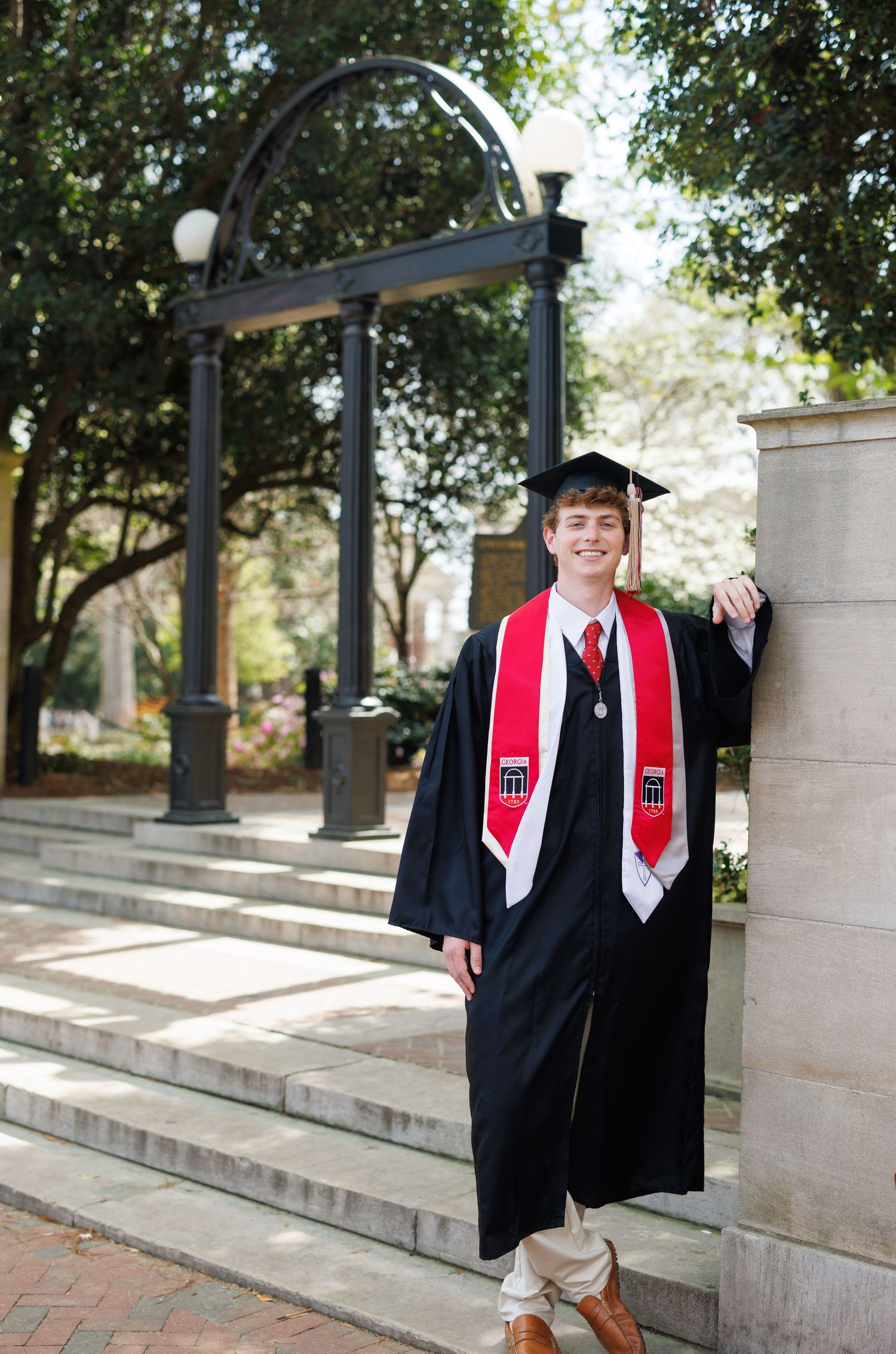 Graduate in black cap and gown with red stole poses by iconic arch on college campus.