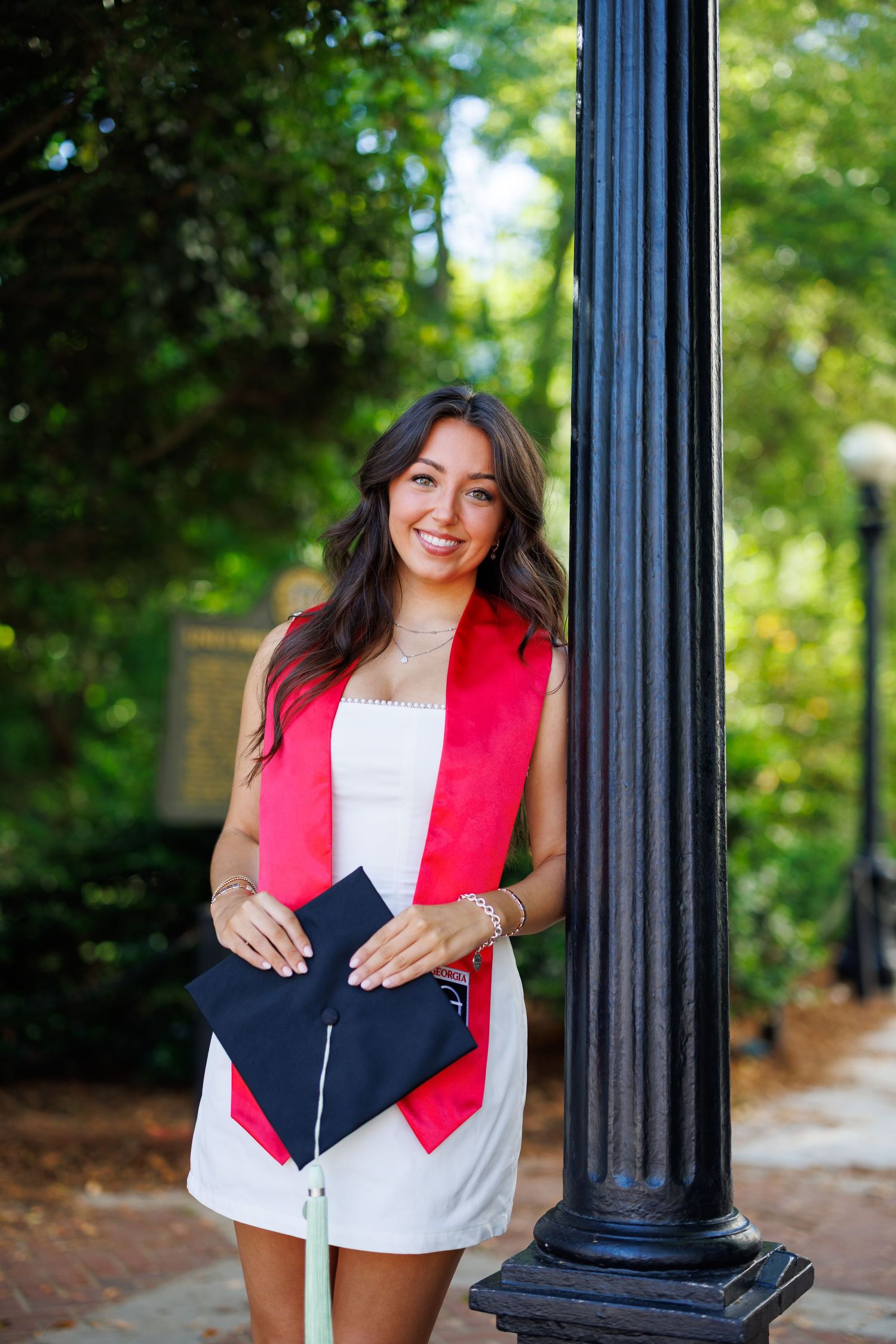 Smiling female graduate in white dress with red stole holds cap near lamppost outdoors.
