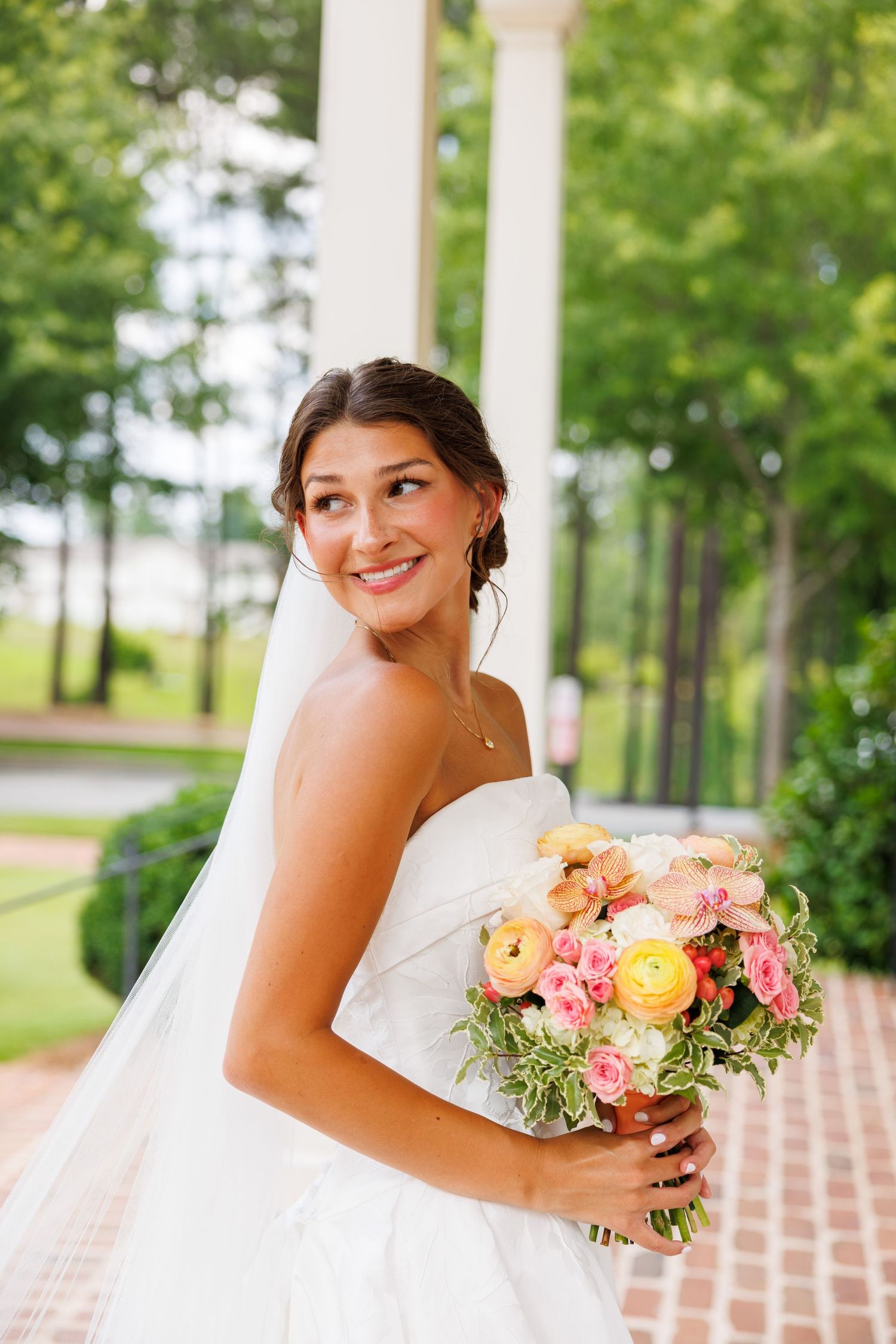 Smiling bride in strapless white gown holding colorful bouquet of pink and orange flowers outdoors near white columns.