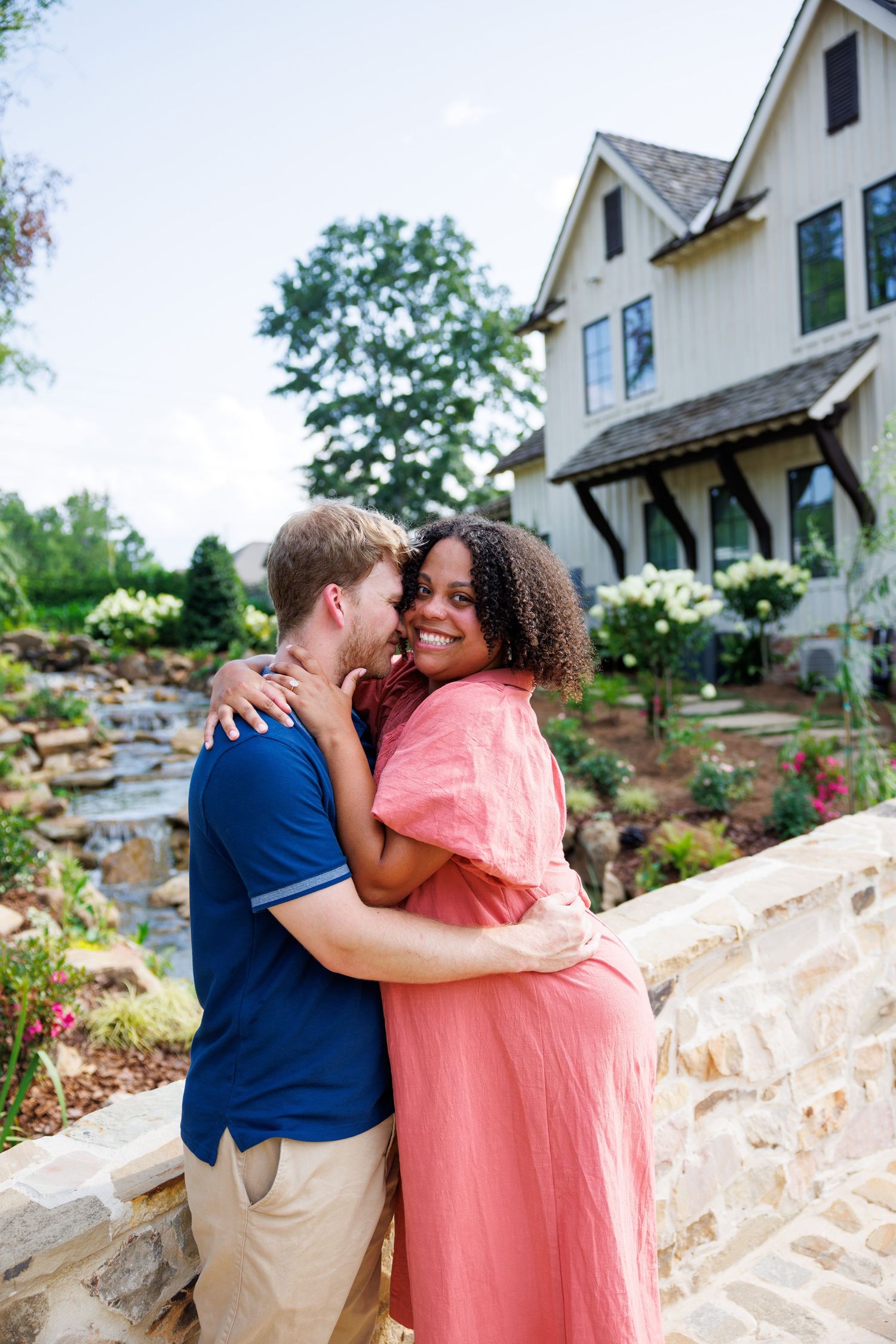 A couple embraces outdoors near a stone wall, woman in pink dress smiling, with a charming farmhouse and waterfall in background.