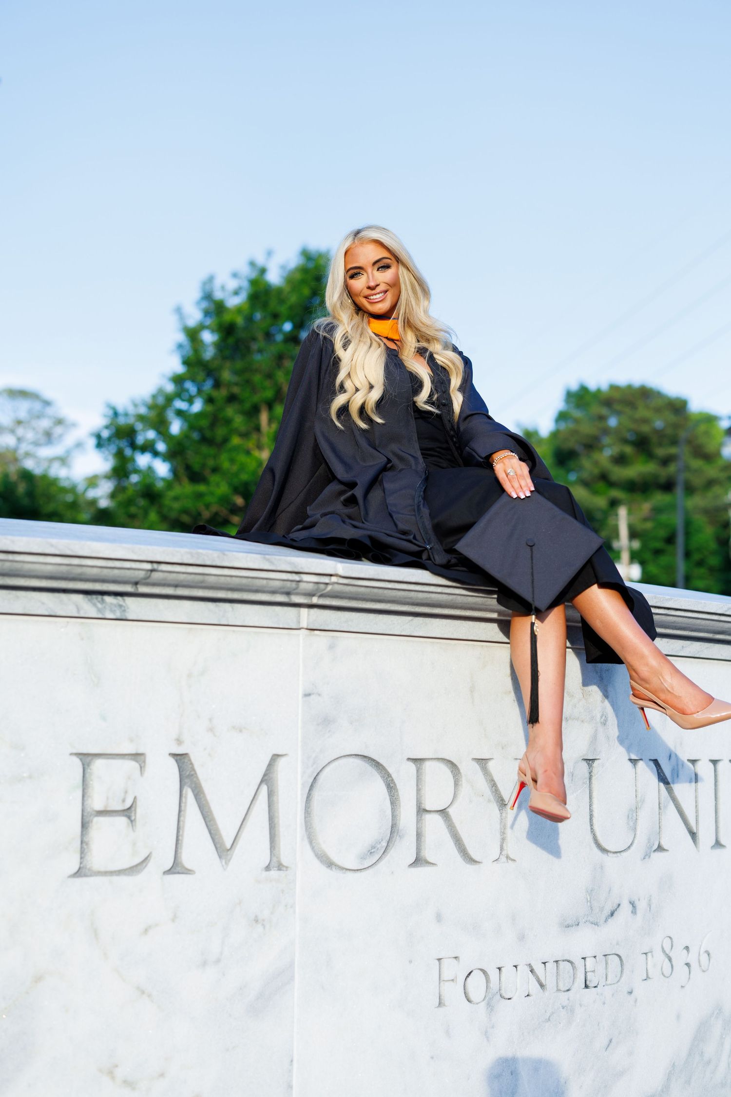 Graduate in cap and gown sits on Emory University stone entrance sign, smiling outdoors.